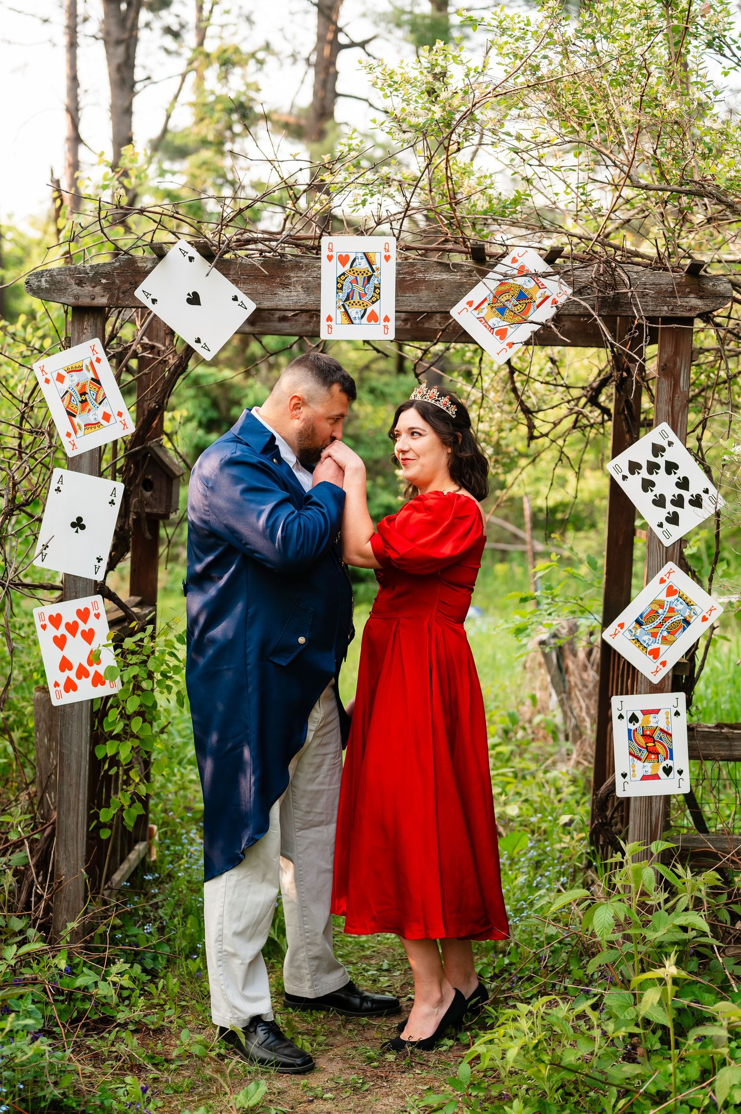 Whimsical couple poses by rustic fence decorated with playing cards in outdoor garden setting.