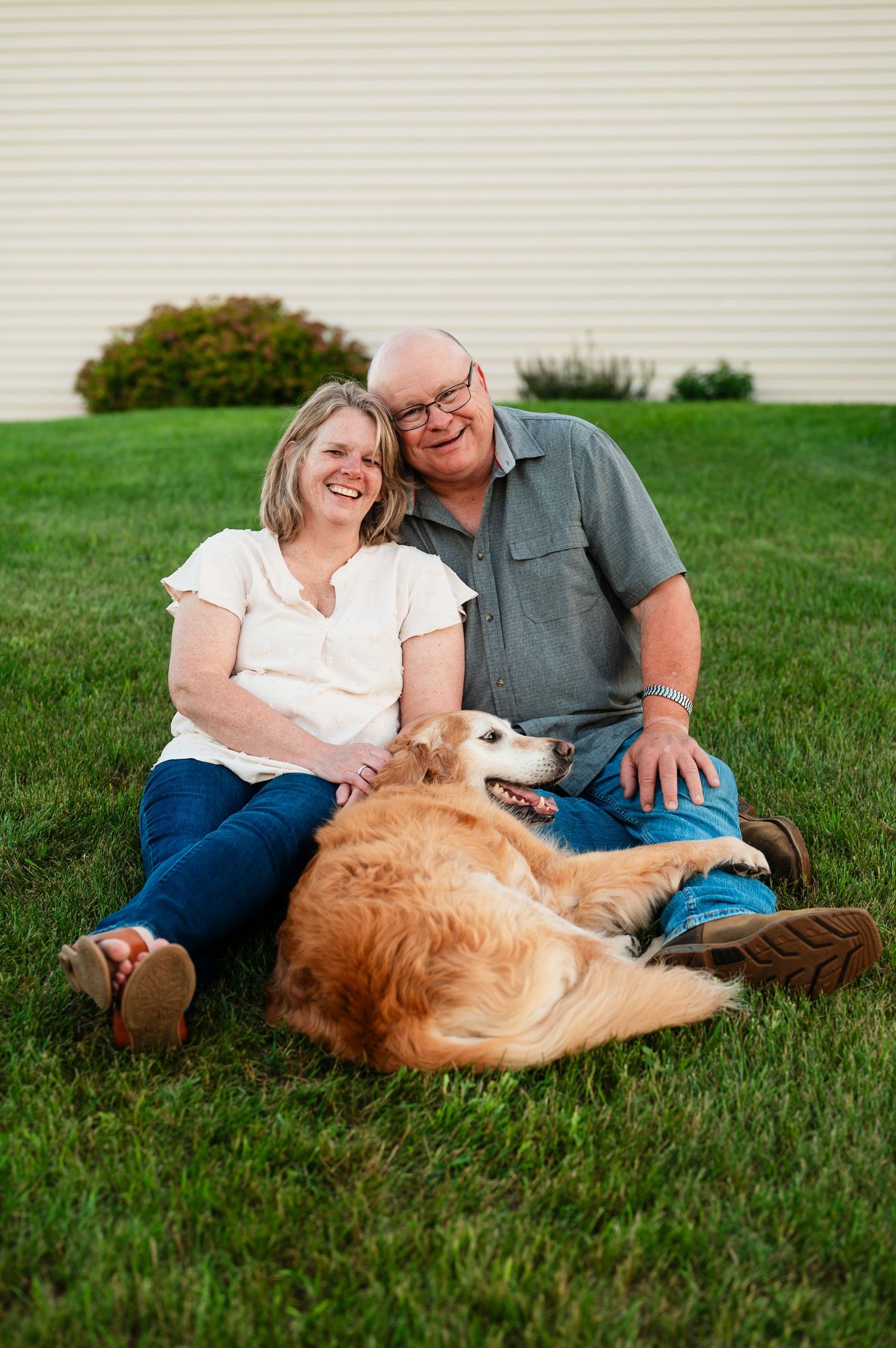 Husband and wife relaxing on grass with their golden retriever dog during outdoor family session.