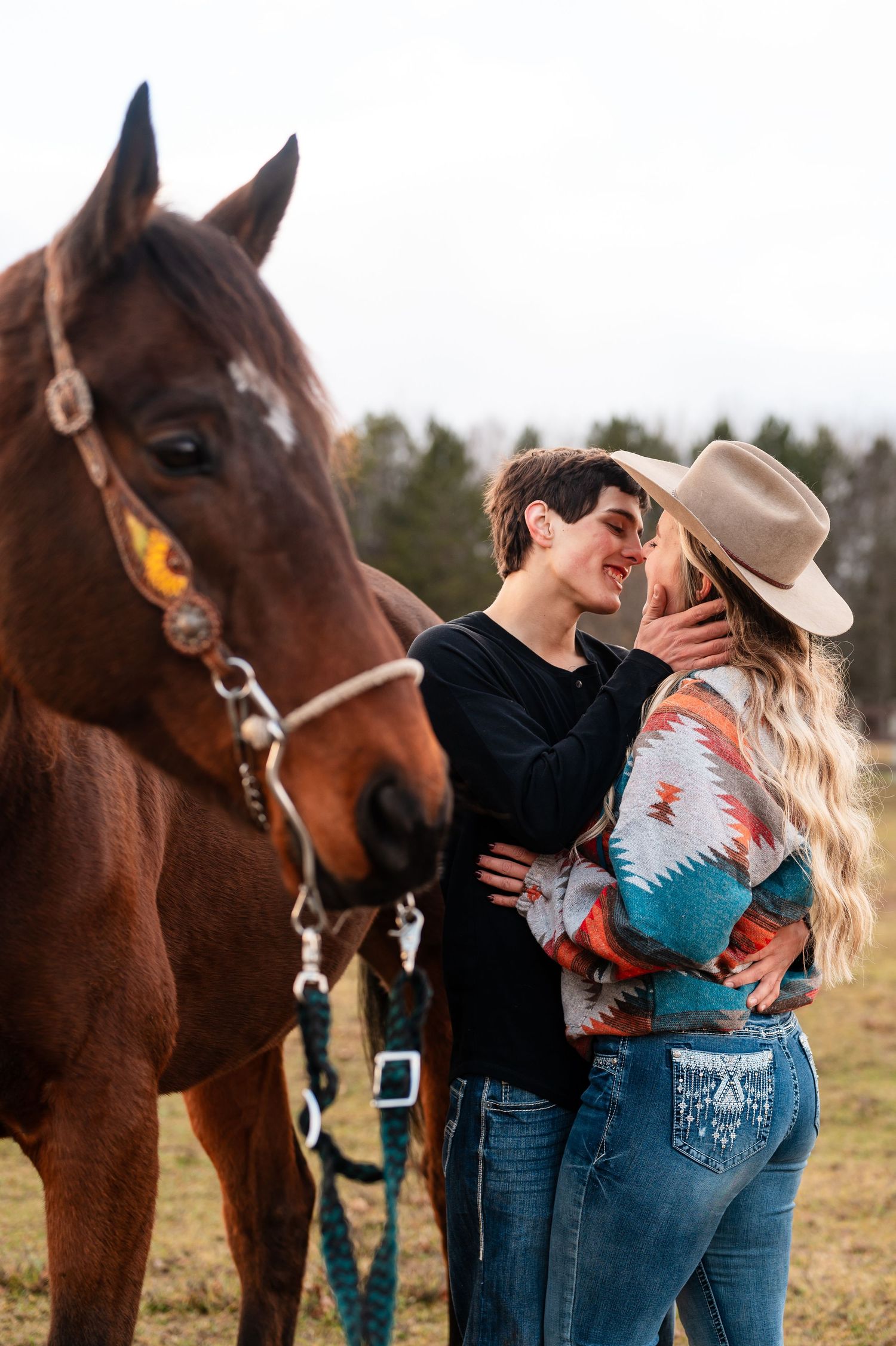 Romantic couple in western wear embraces near a brown horse in a sunny field.