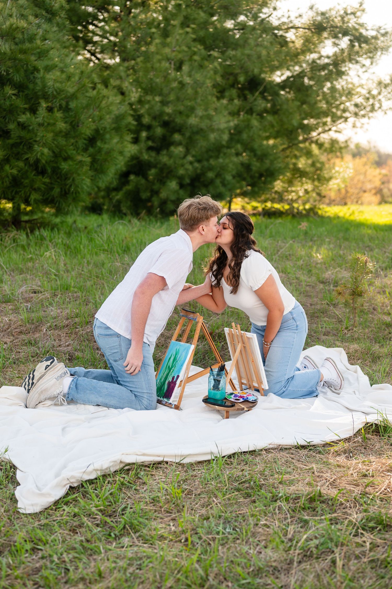 A couple enjoys a painting session on a white blanket in a grassy outdoor setting.