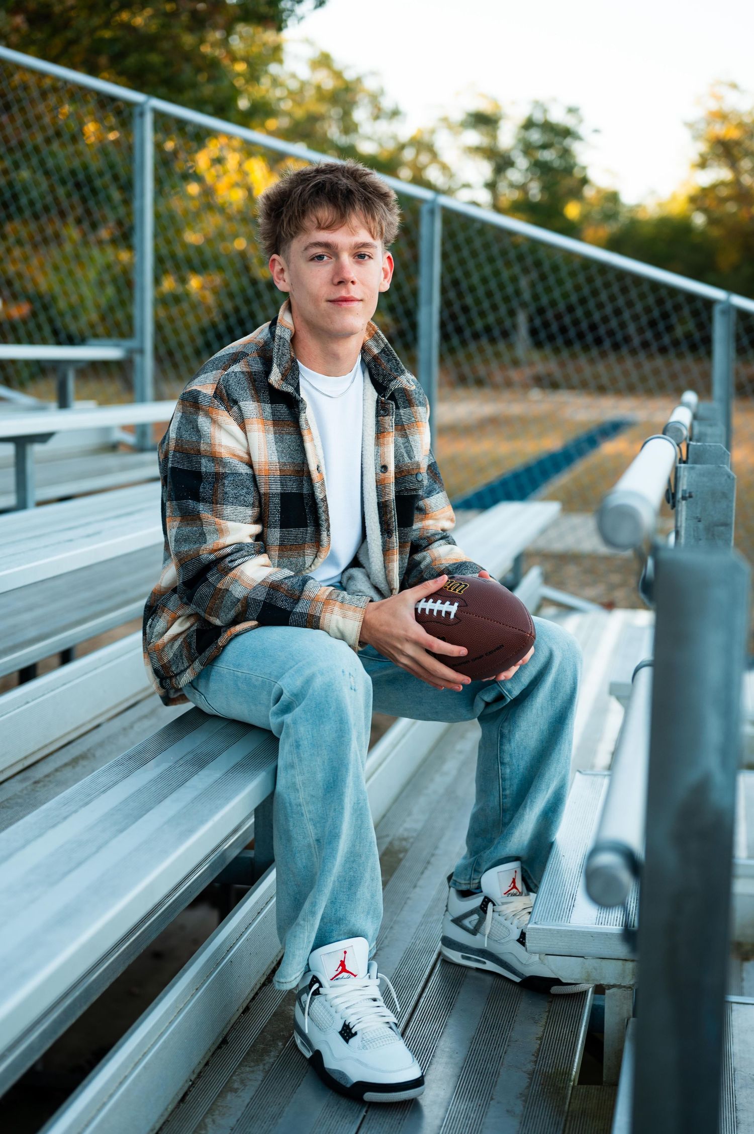 Senior boy on metal bleachers holding football wearing plaid shirt and jeans.