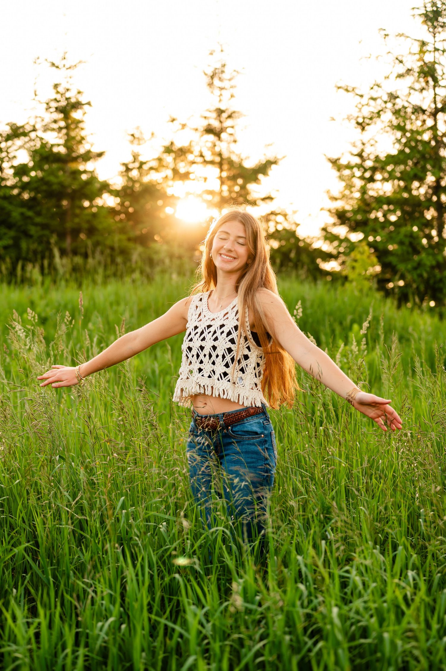 A carefree senior girl dances joyfully through a sunlit field wearing a patterned crop top and jeans at golden hour in Northern Michigan.