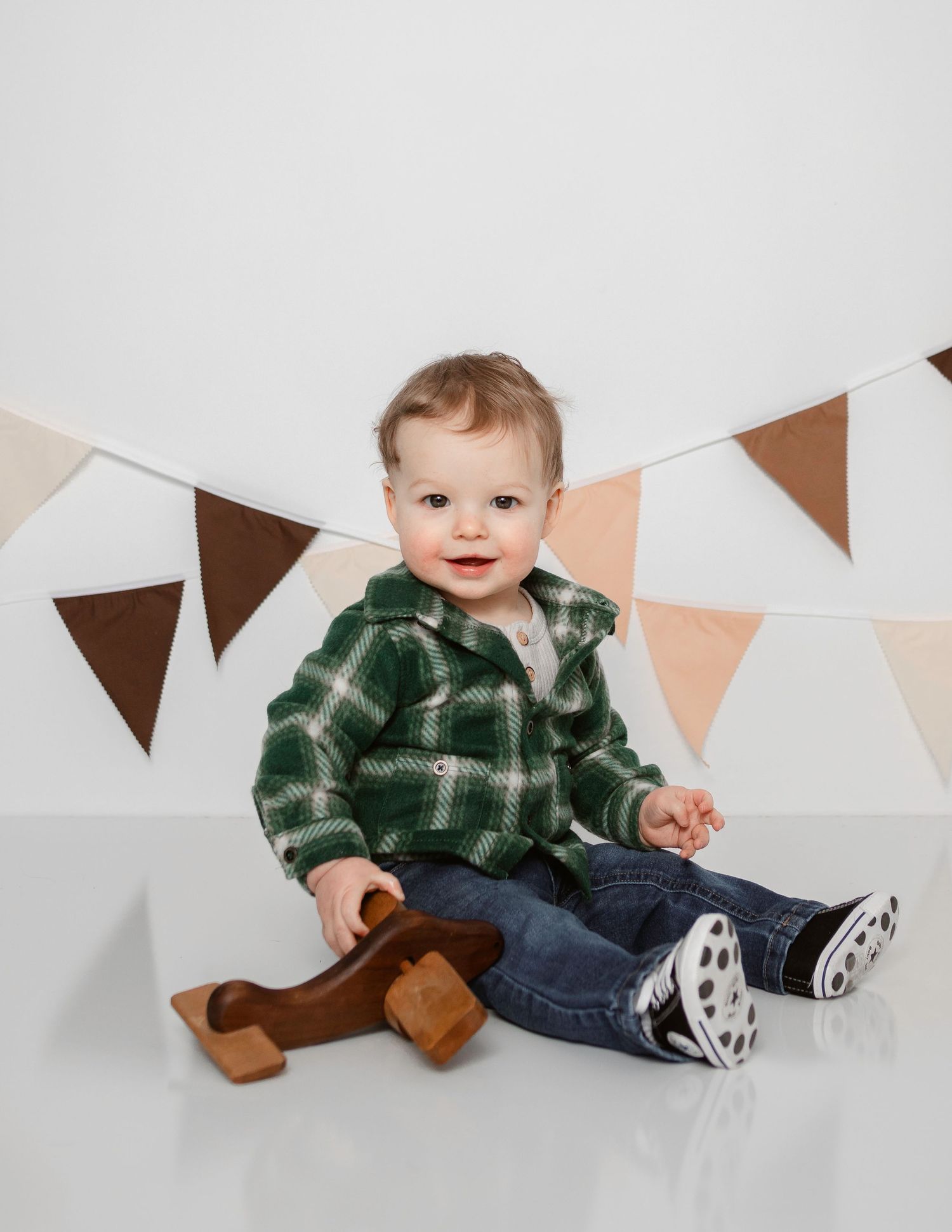 Baby in green plaid shirt and jeans sitting against white wall with brown and peach bunting.