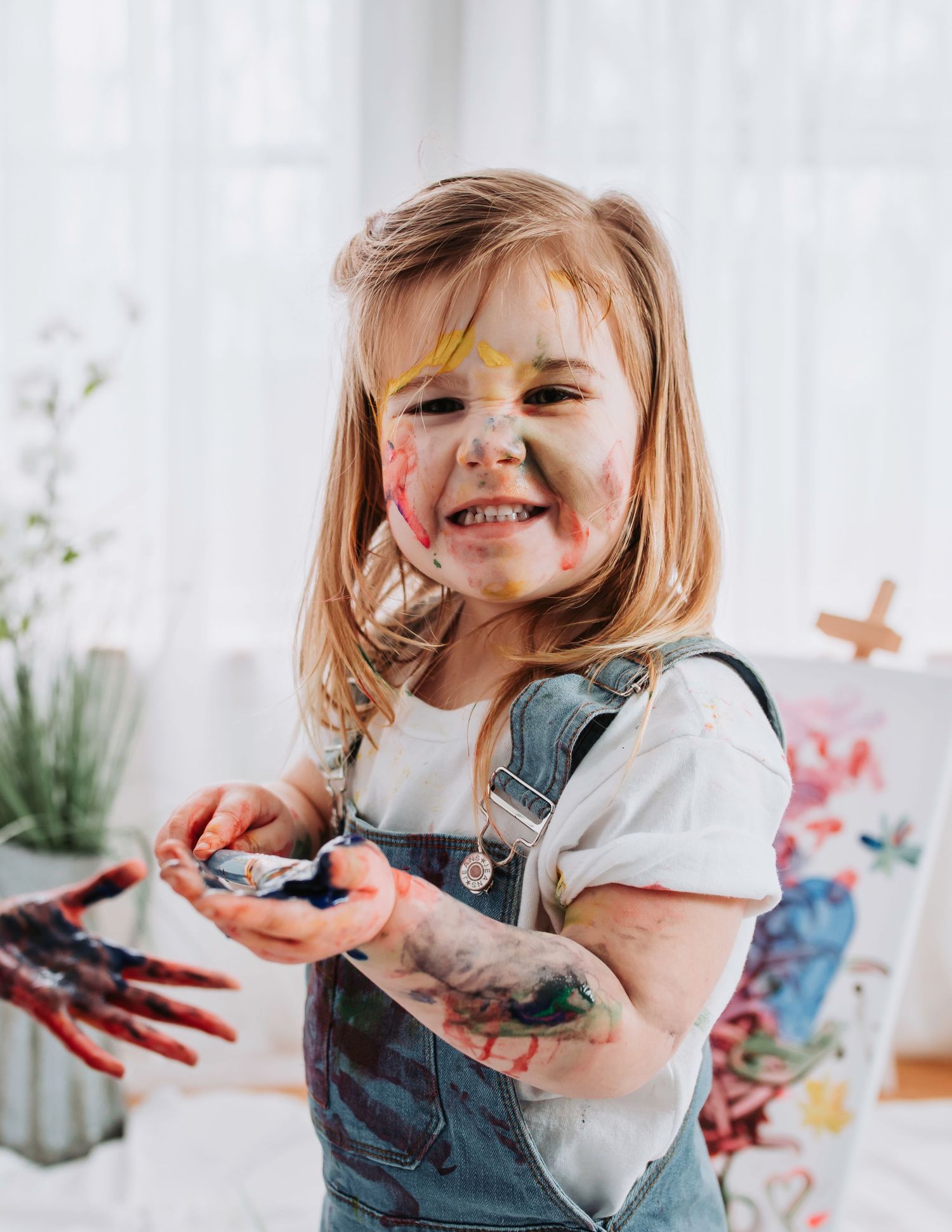 A creative painting session shows colorful paint-stained hands and a bright smile in denim overalls.