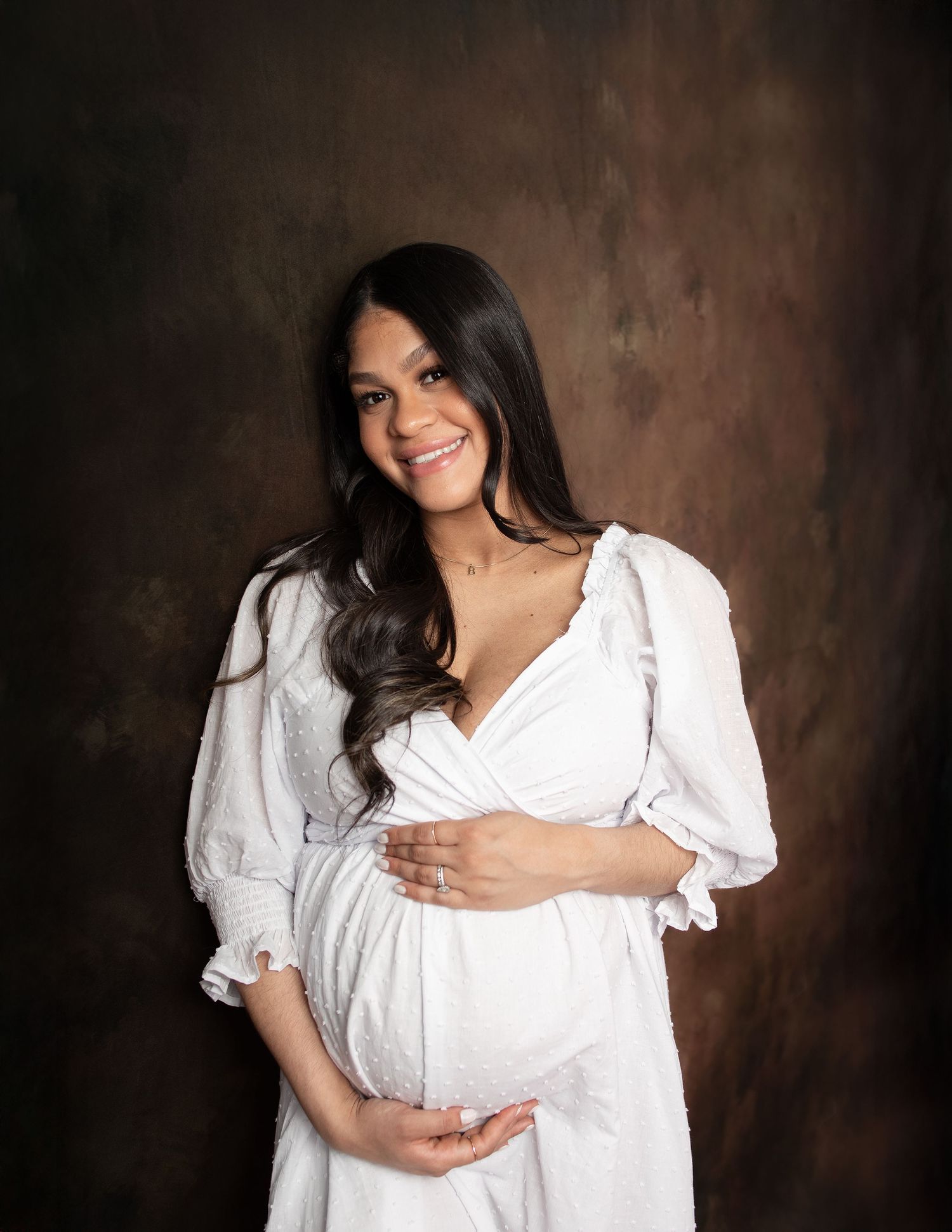 Woman in white dress poses for maternity photos against dark textured backdrop.