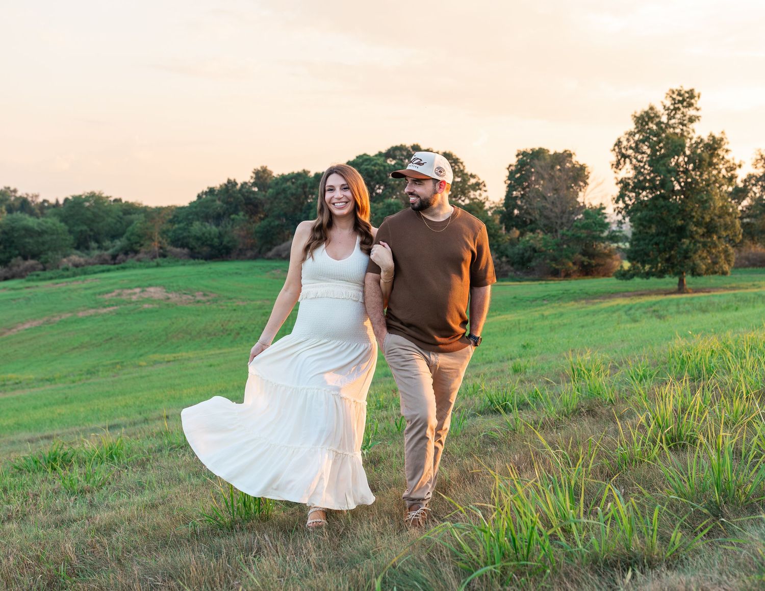 Couple walking hand-in-hand through a lush green field at sunset, woman in white dress, man in brown shirt.