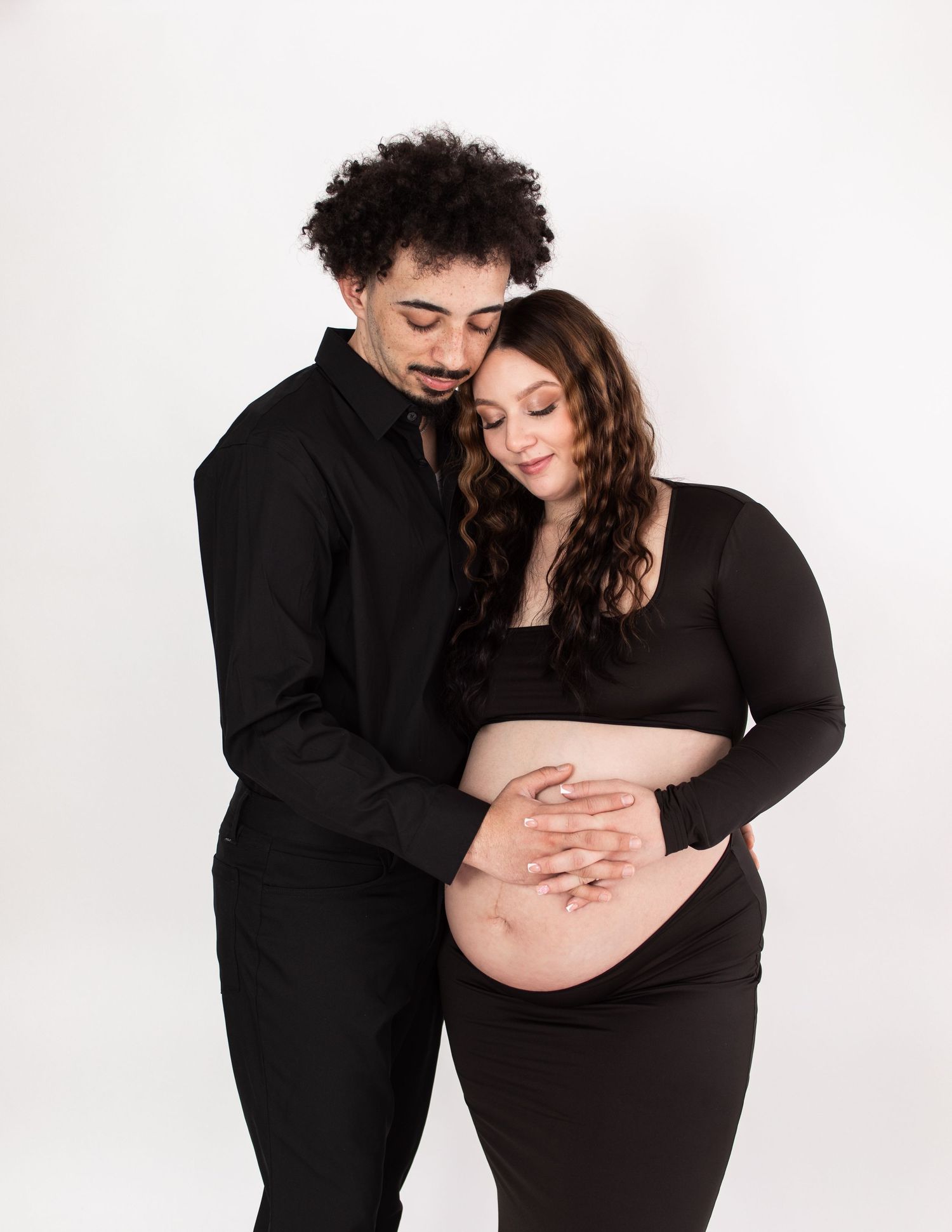 Intimate maternity photos of couple wearing black outfits against white backdrop.
