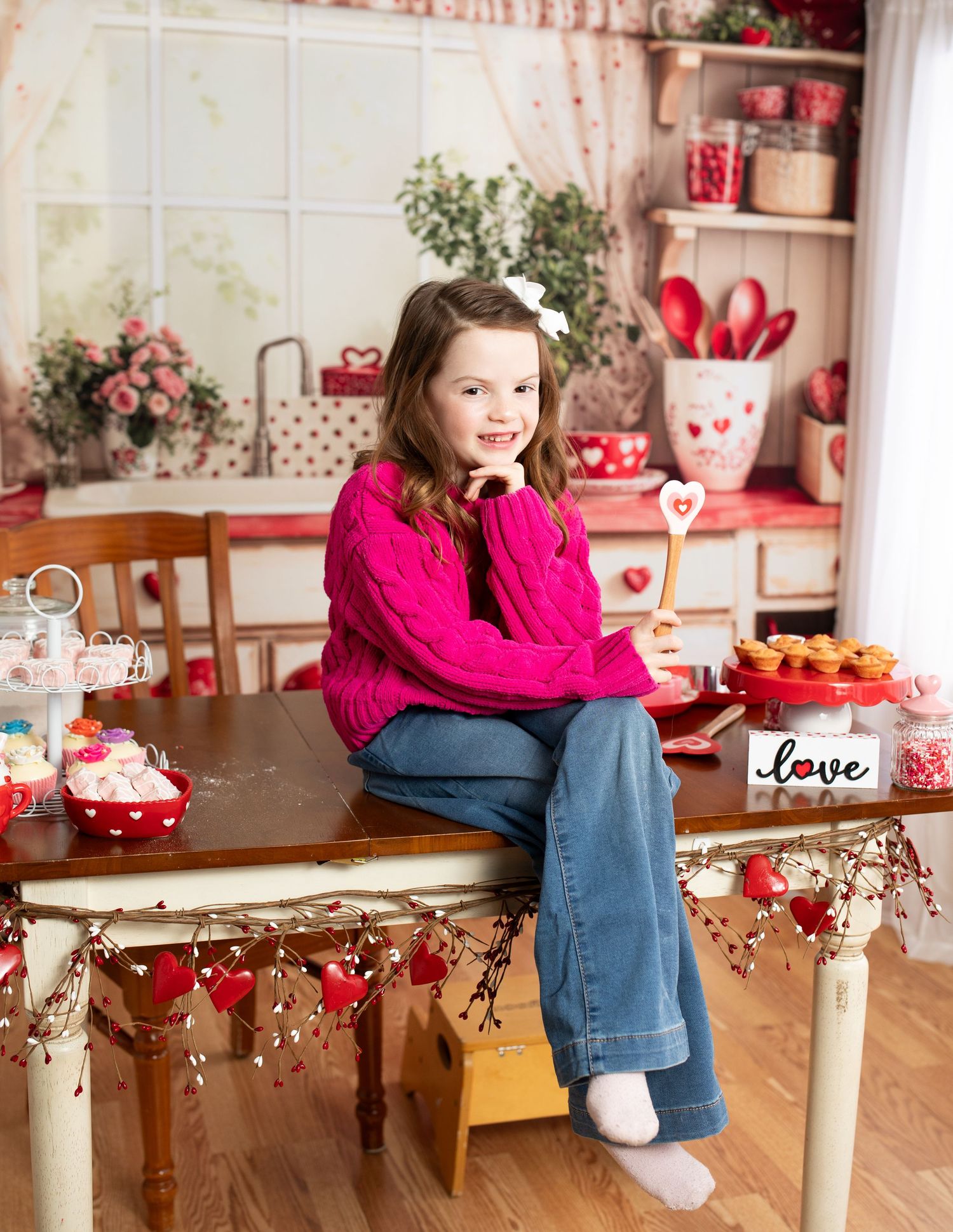 Girl in pink sweater sitting among Valentine's Day decorations and heart-themed kitchen items.