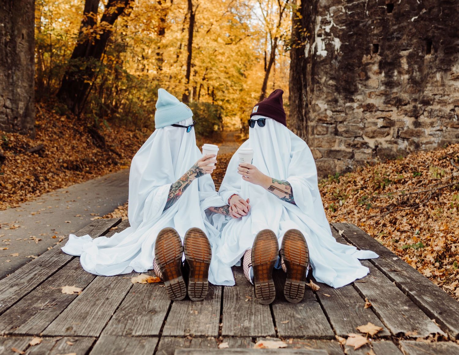 Two figures in ghost costumes lounging on wooden planks surrounded by fall leaves.