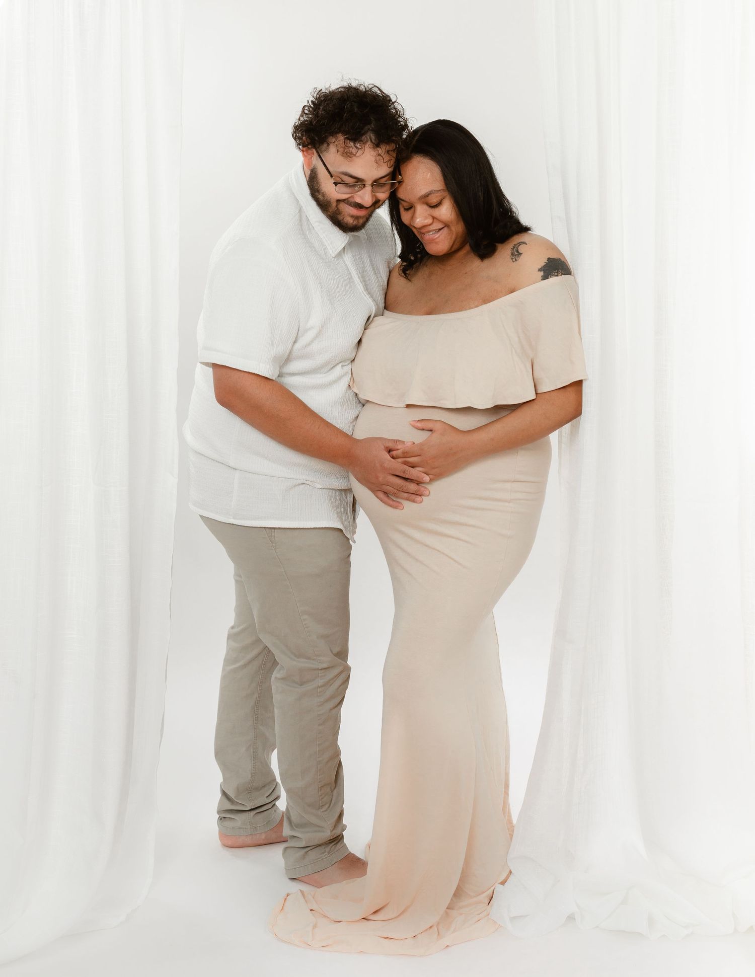A couple in white and beige clothing shares intimate maternity portraits against a light backdrop.
