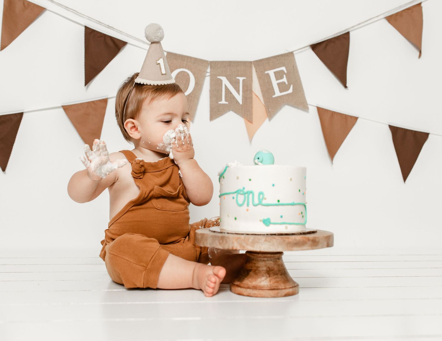 Baby in brown outfit sits on white backdrop with ONE banner and cake during first birthday cake smash photo session.