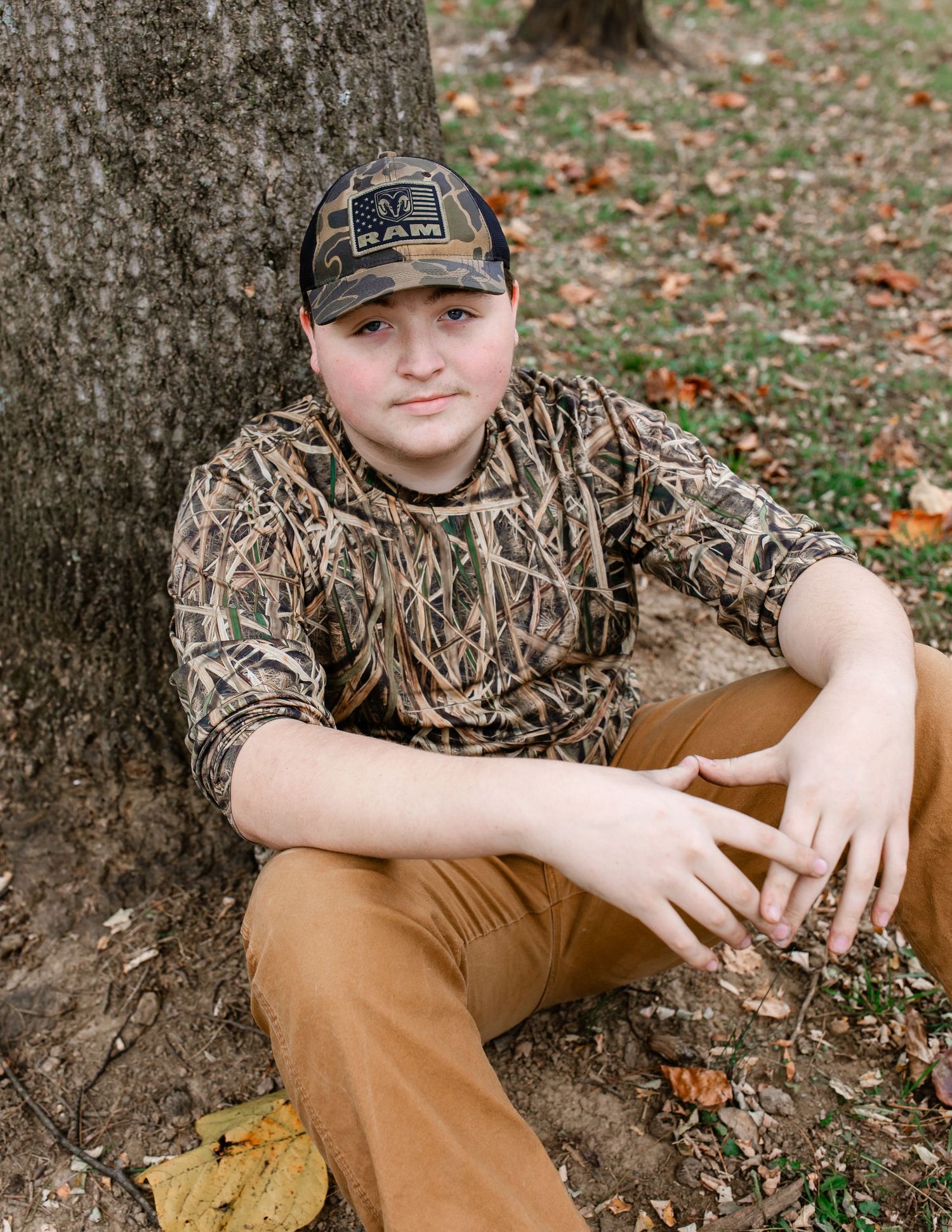 Young person in camo shirt and brown pants sits against large tree trunk in autumn setting.