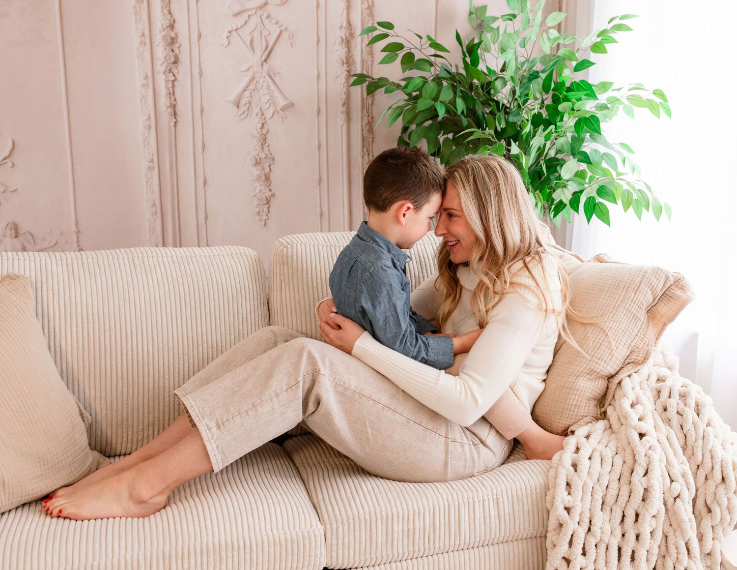 A mother and young son share a tender moment on a cream sofa with a green plant and chunky knit blanket.