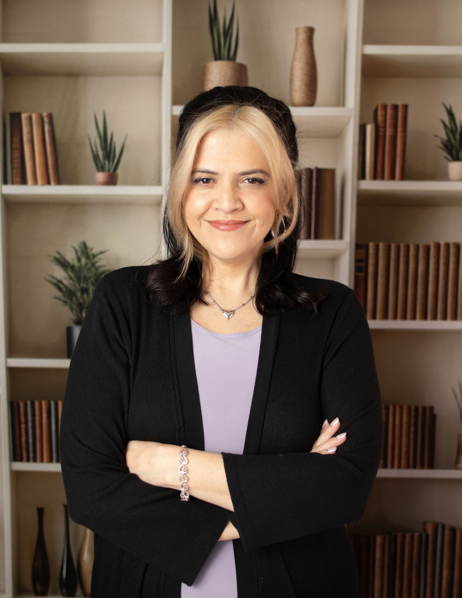 Professional wearing a black blazer and purple top poses with crossed arms against white built-in bookshelves.