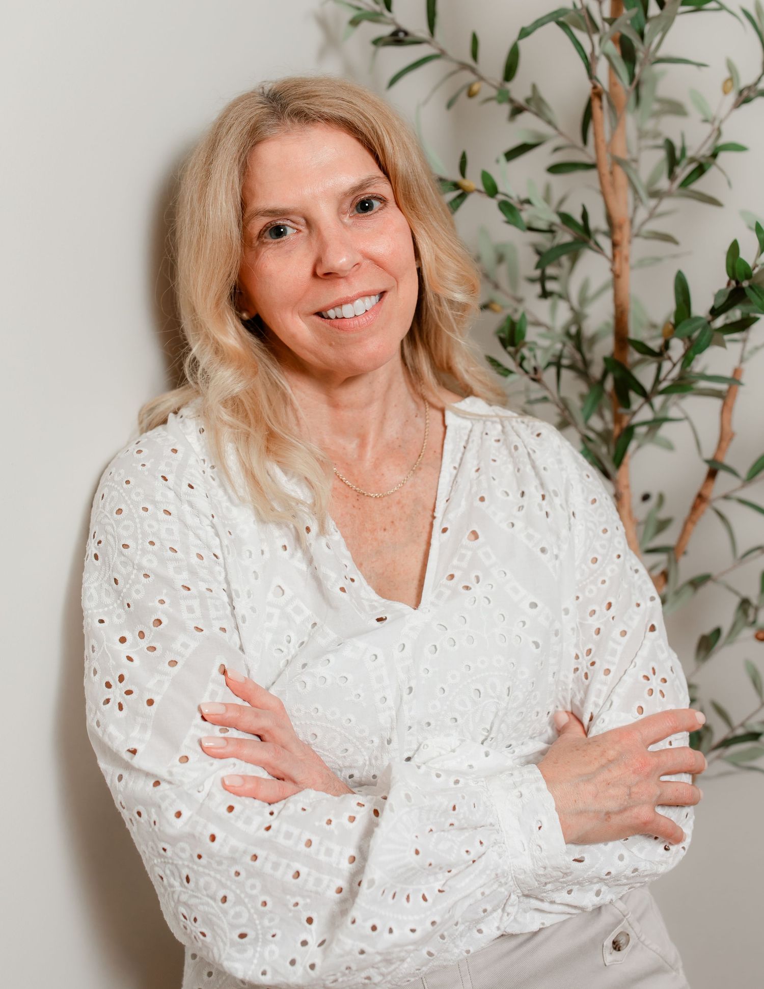 Person in white eyelet blouse posing against plant backdrop.