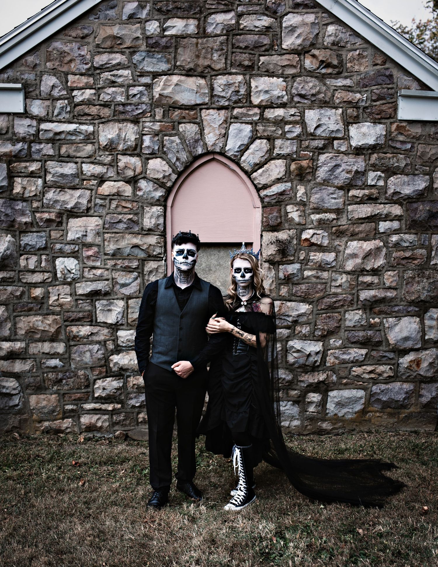Gothic couple with skull makeup poses against stone building wall.
