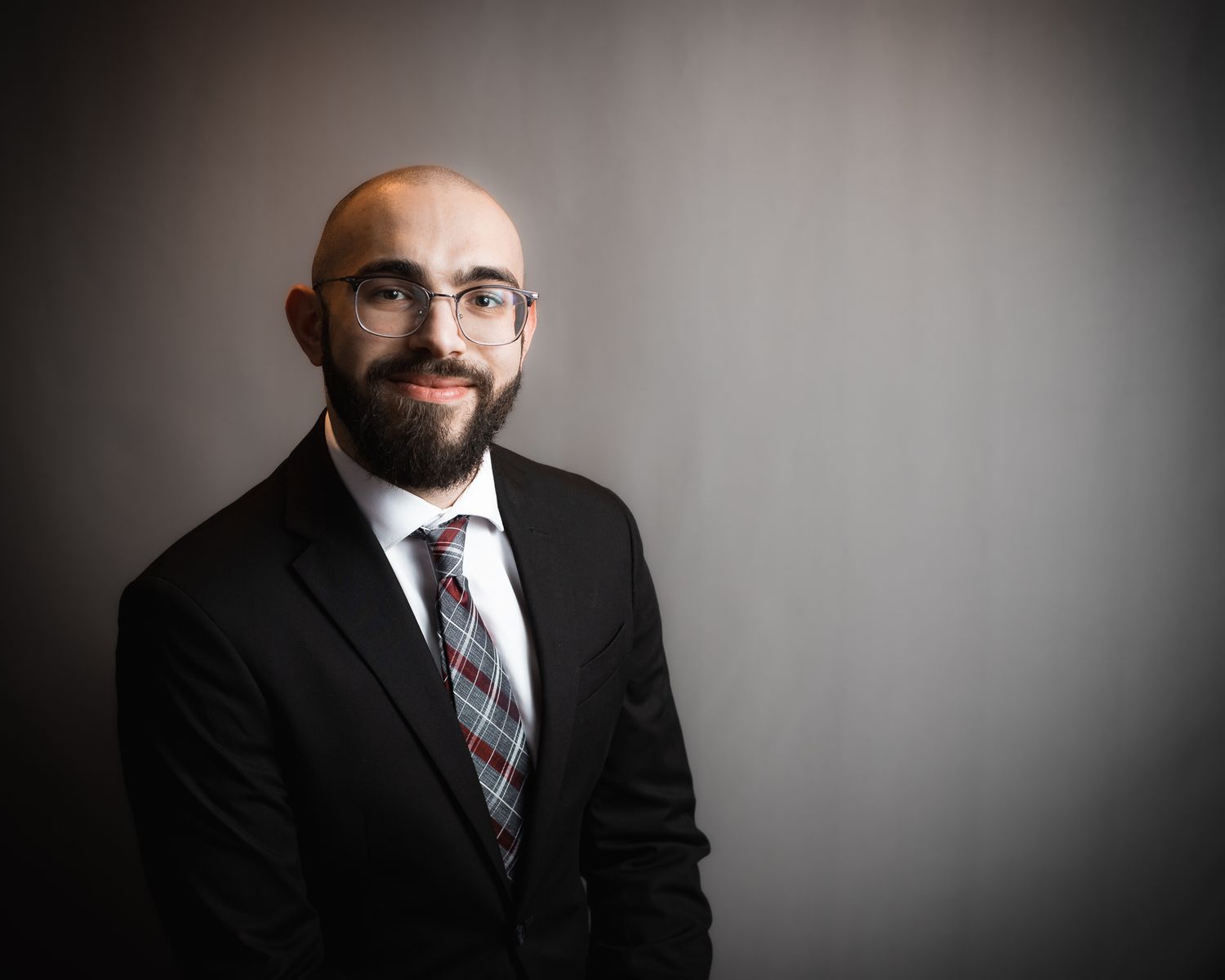 Professional headshot of a bearded man in a dark suit and plaid tie against a gray background.