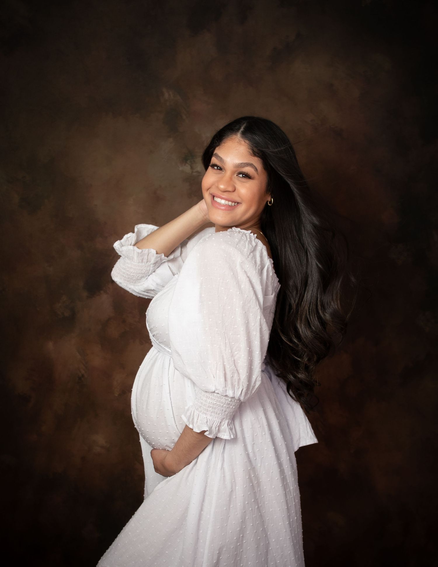 Joyful maternity portraits of woman in flowing white dress against dark backdrop.