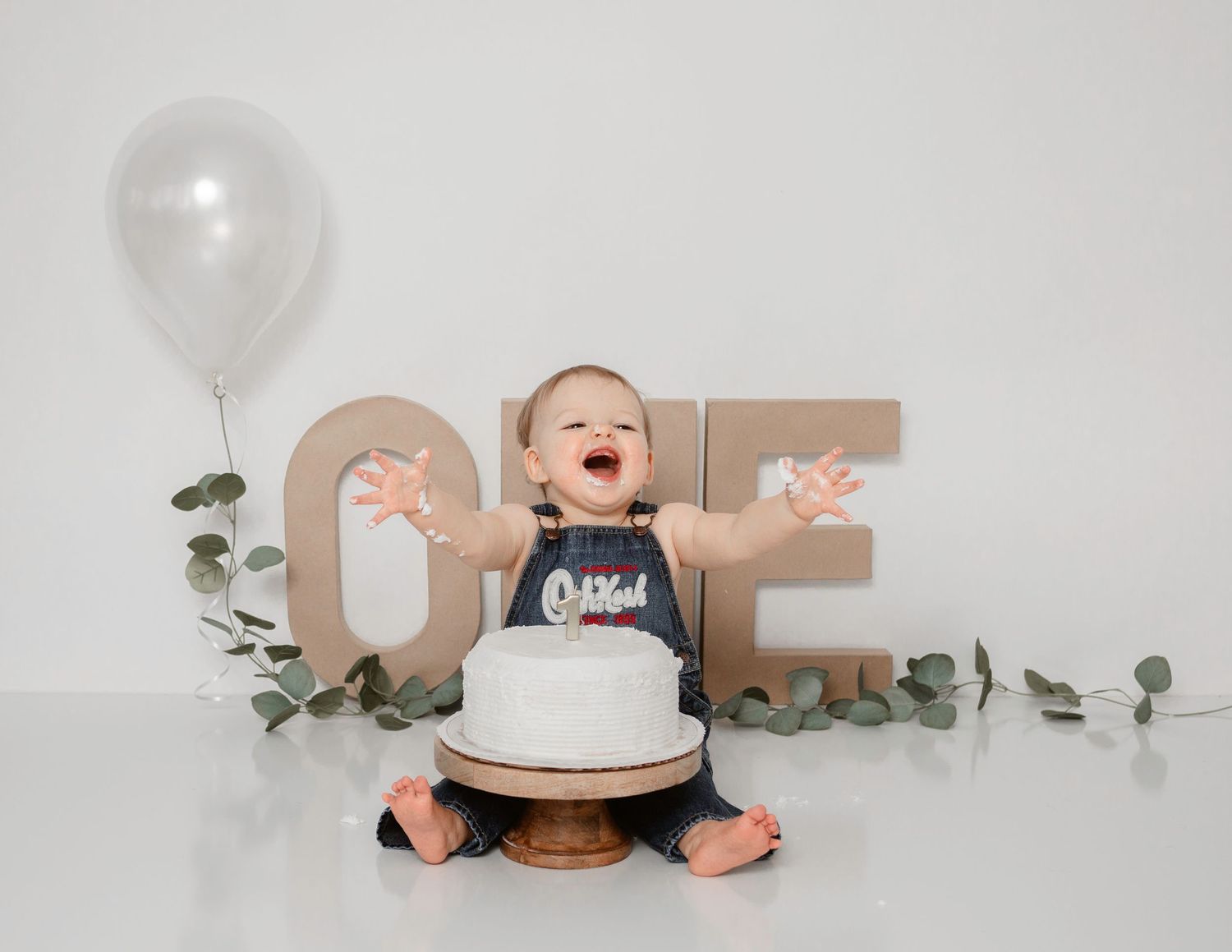 Baby reaching for white birthday cake next to wooden ONE letters and balloon decor.