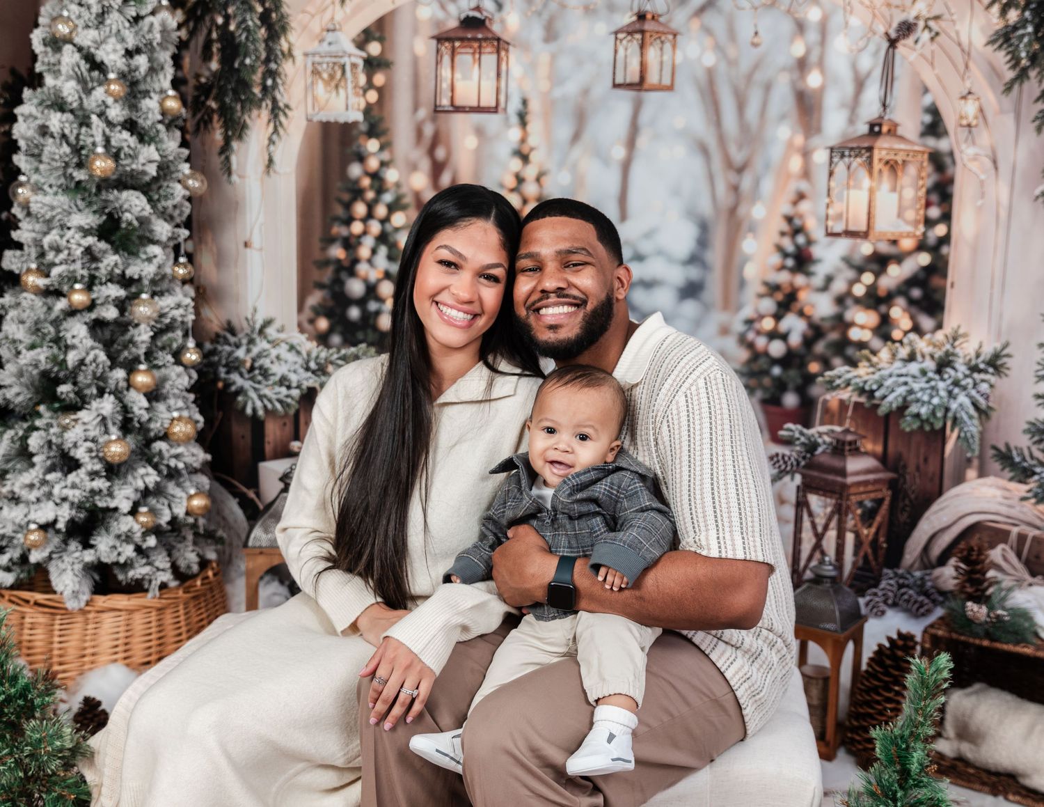 Family poses together in cozy winter setting with frosted Christmas trees and lantern decorations in background.