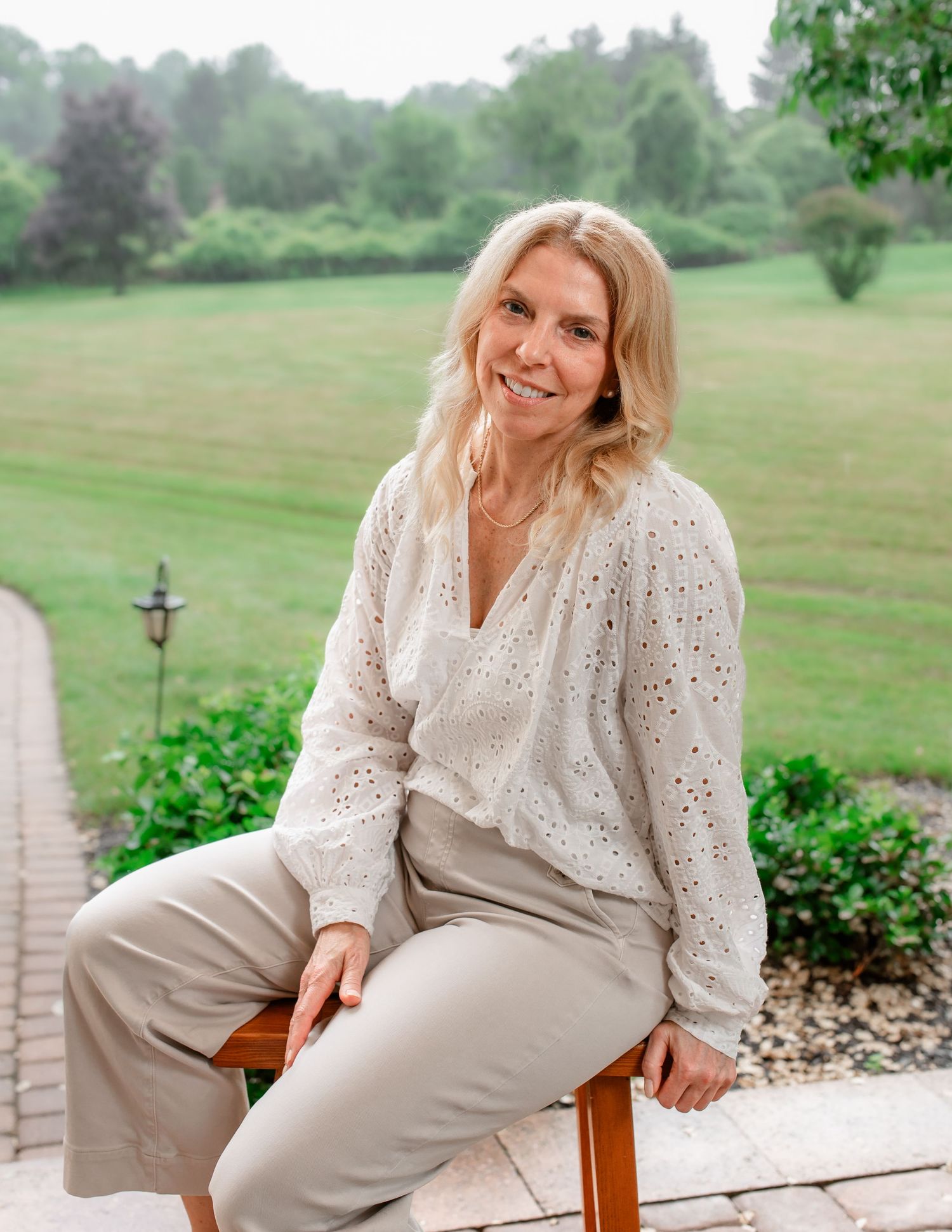 Person in white eyelet top and beige pants sitting outdoors.
