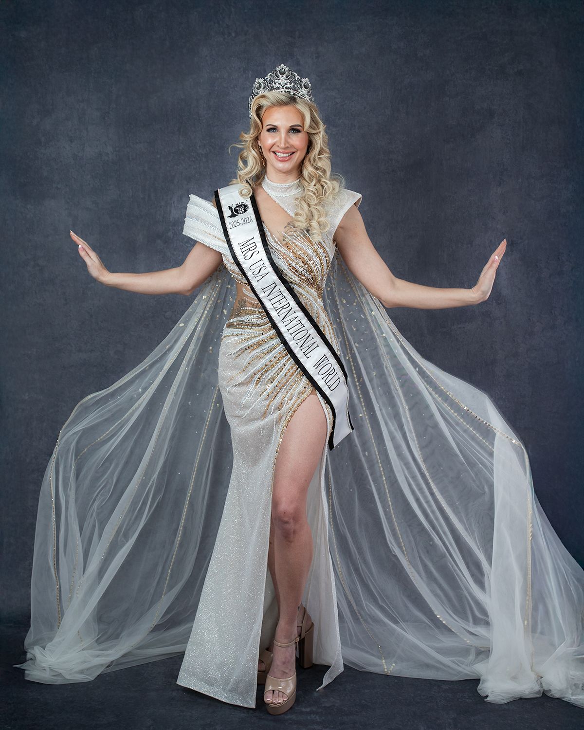 Pageant contestant in flowing silver gown and tiara poses with arms outstretched.