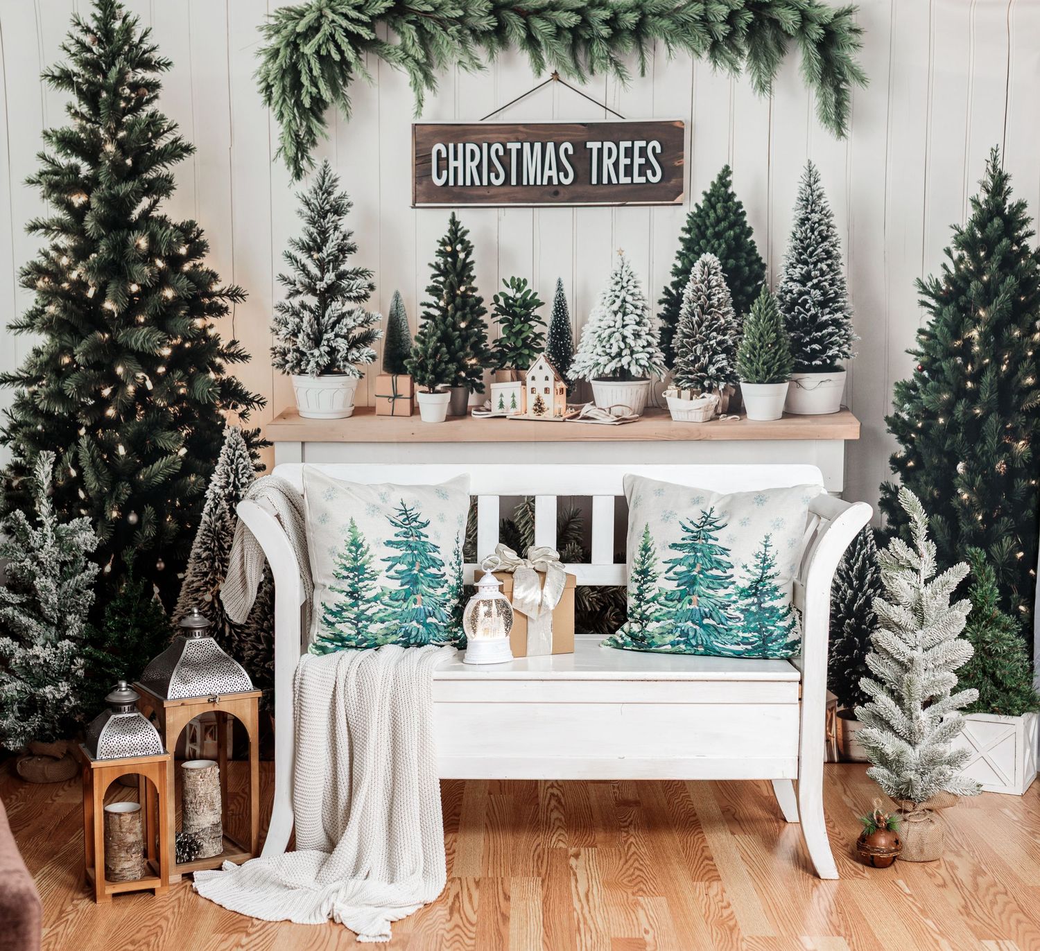 Festive Christmas vignette with white bench, decorated trees, pine pillows, lanterns, and a Christmas Trees sign.