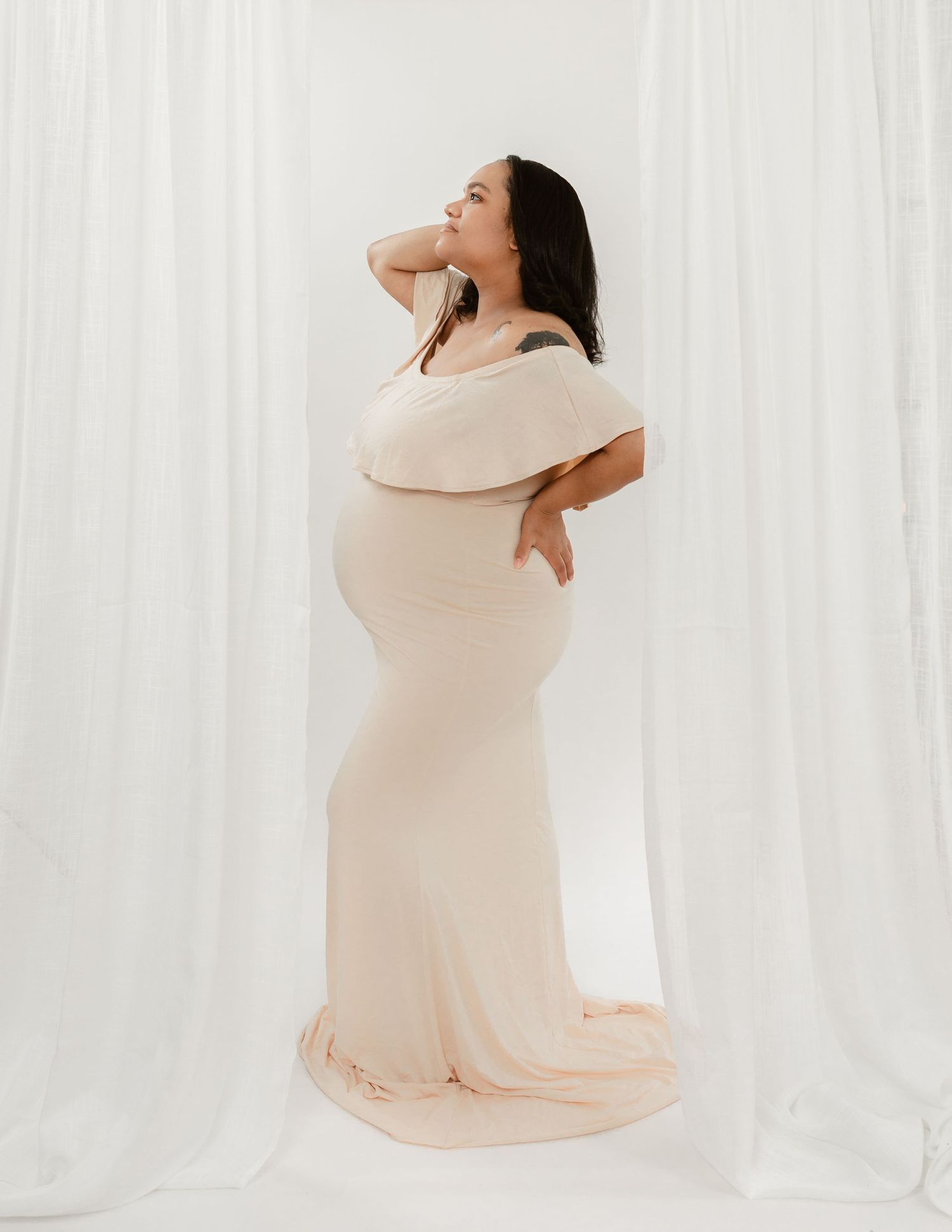 A pregnant woman in a cream dress poses gracefully against white curtains in a maternity photoshoot series.