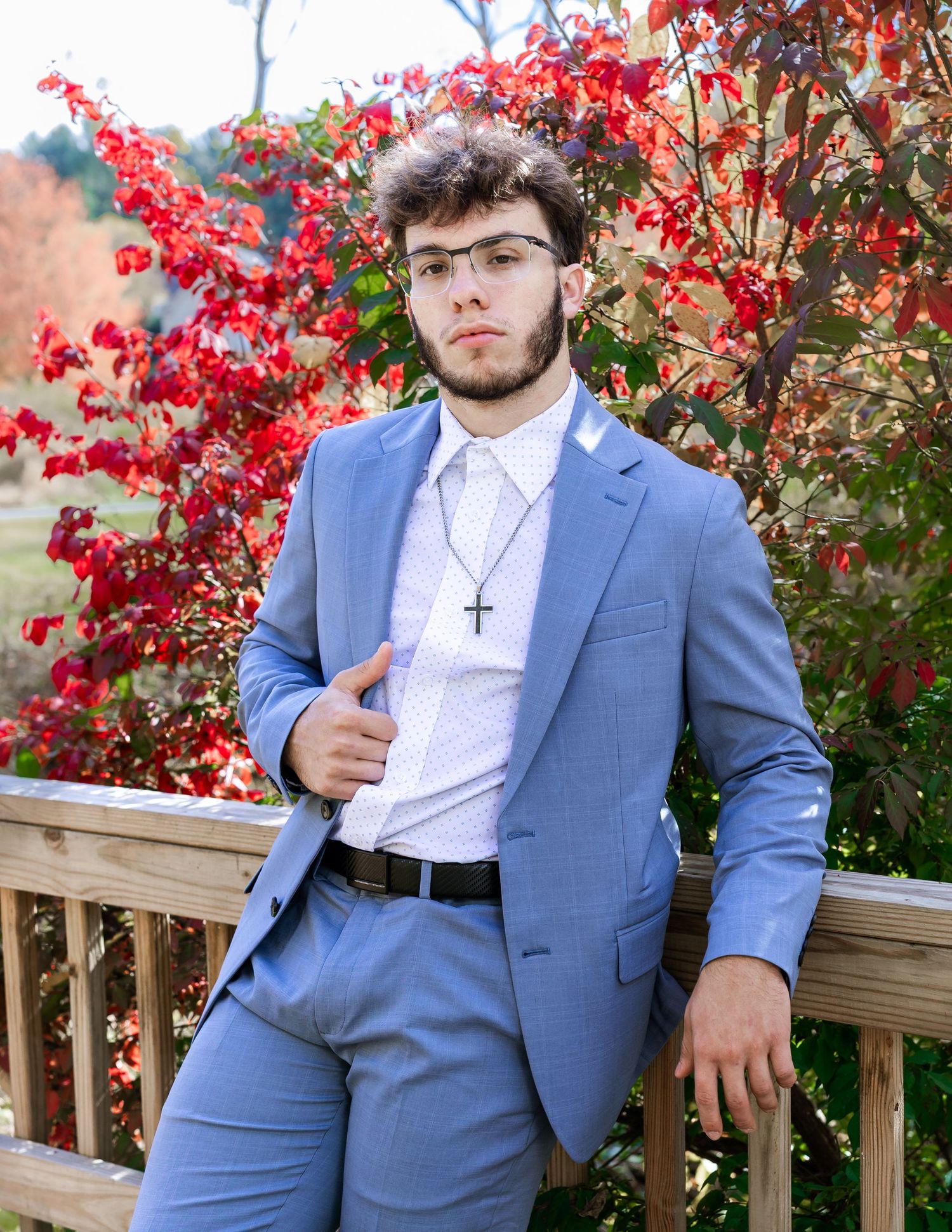Person in light blue suit and white shirt poses against vibrant red autumn foliage.