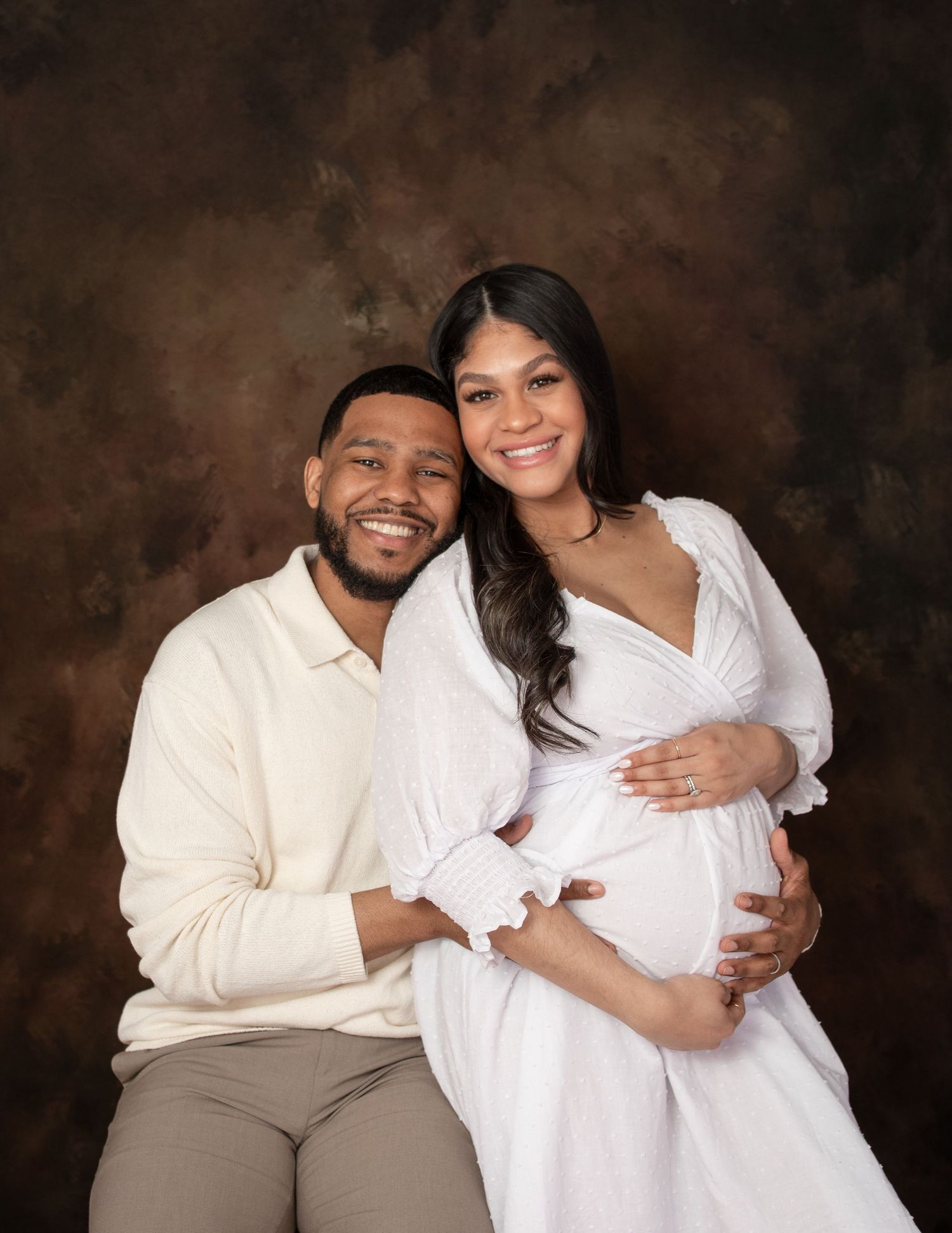A maternity photoshoot featuring a couple in white outfits against a dark brown backdrop.