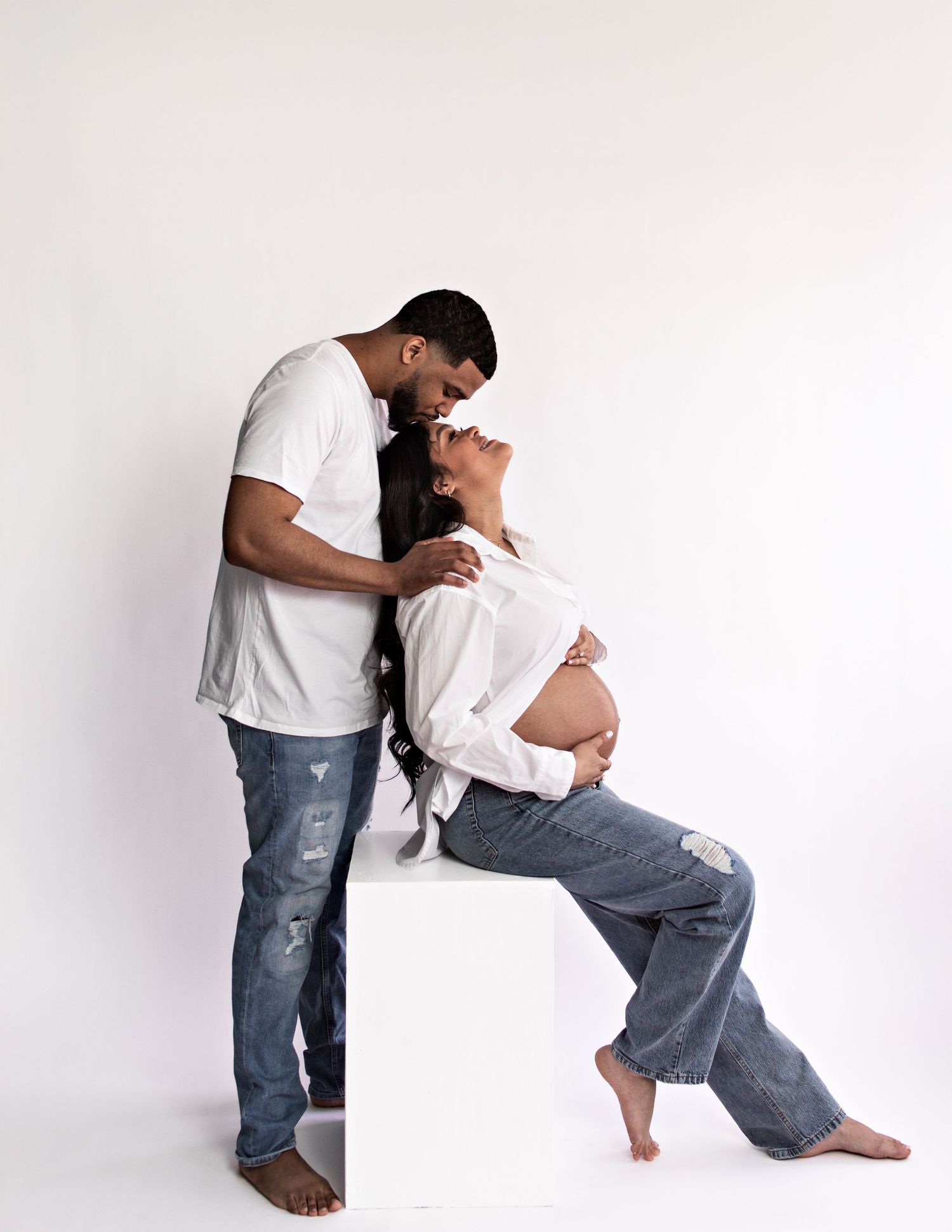 Artistic maternity photos of couple in casual wear against white backdrop.