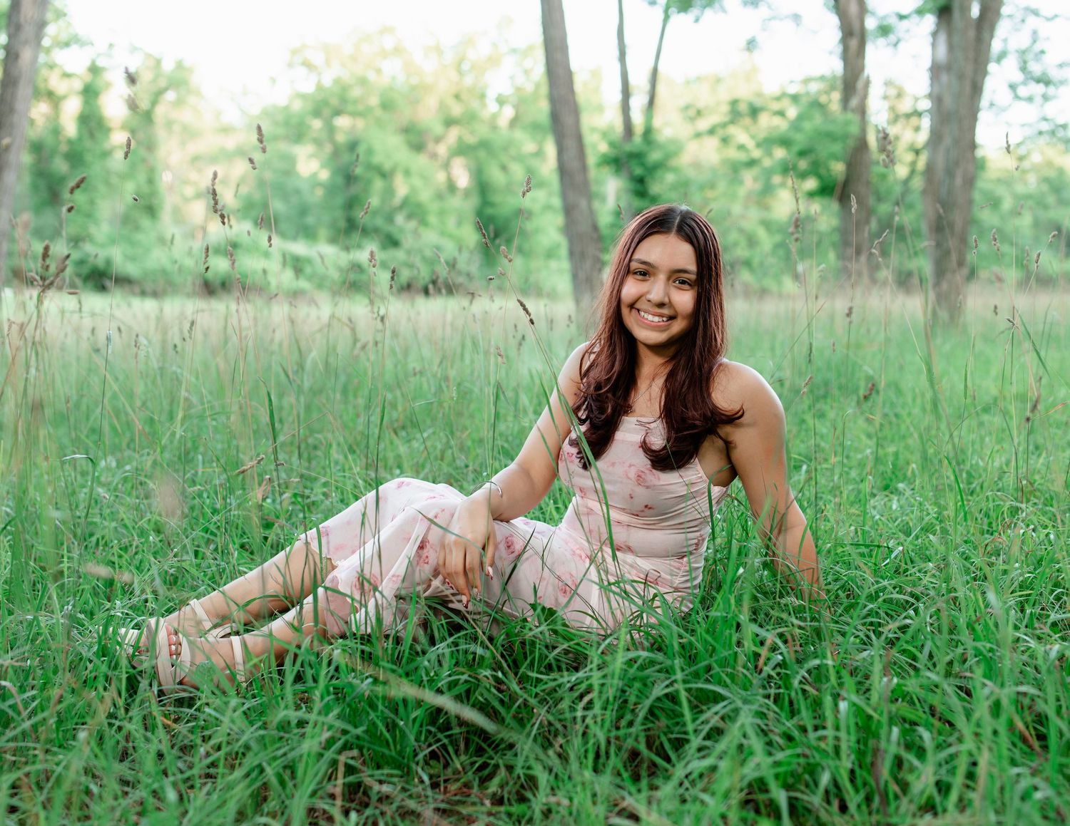 A person in a white dress lies in a grassy field surrounded by trees during a sunny summer day. Senior Session