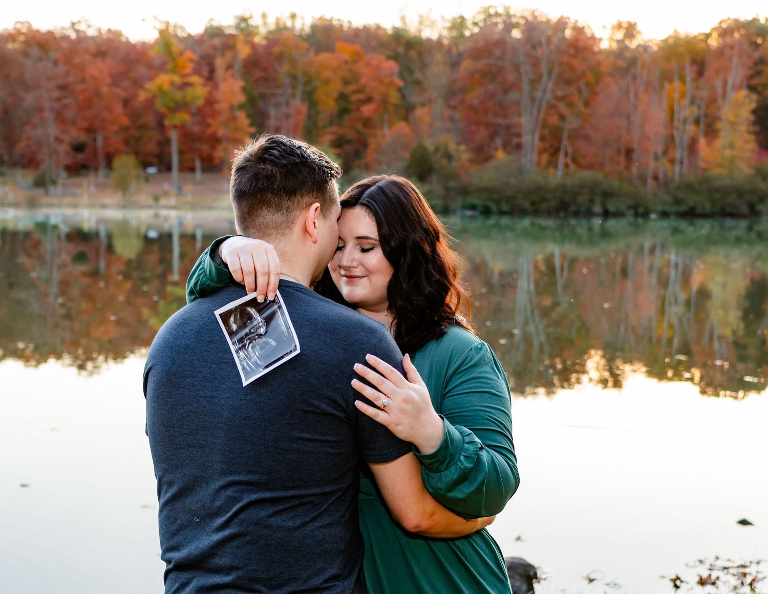 Romantic lakeside maternity photos with couple embracing during autumn sunset.