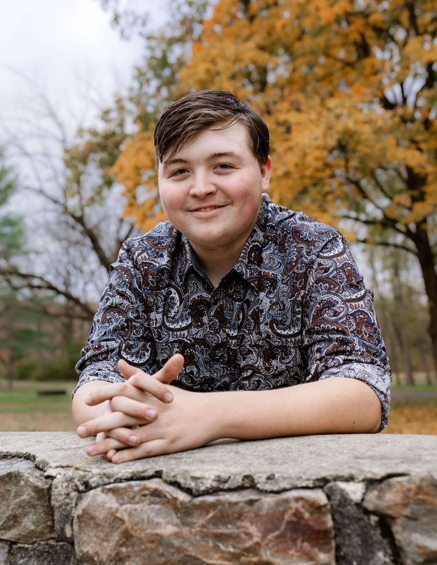 Young person in dark patterned shirt leans on stone wall with autumn foliage in background.