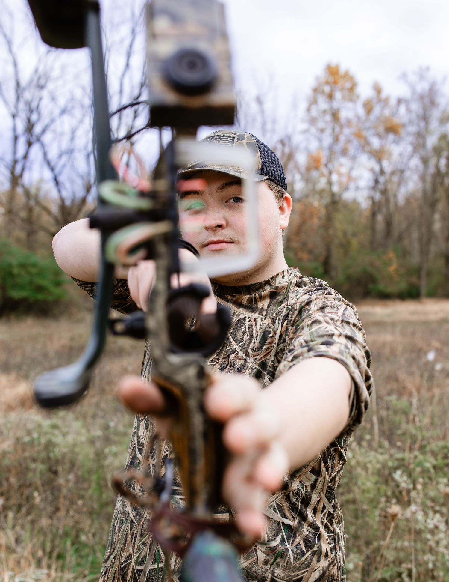 Hunter in camouflage gear aims compound bow in autumn field setting.