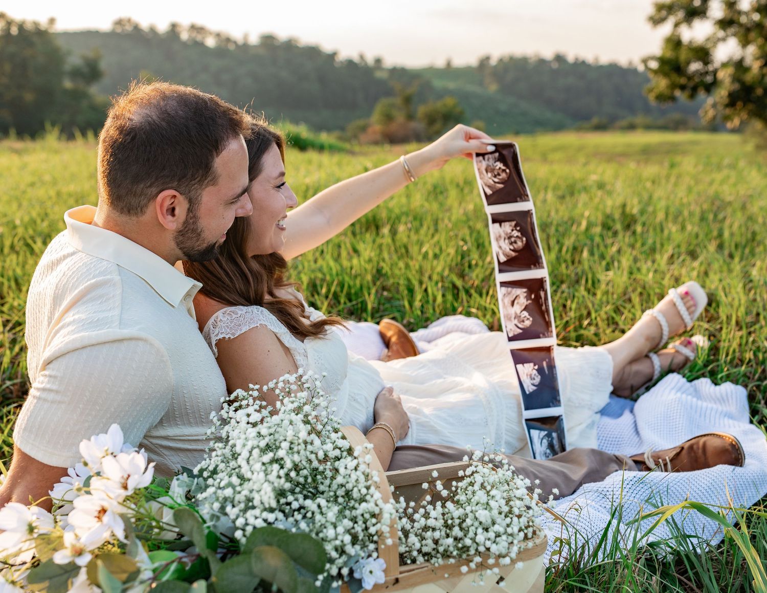 Couple sitting in green field holding ultrasound photos, surrounded by white flowers during golden hour maternity shoot.