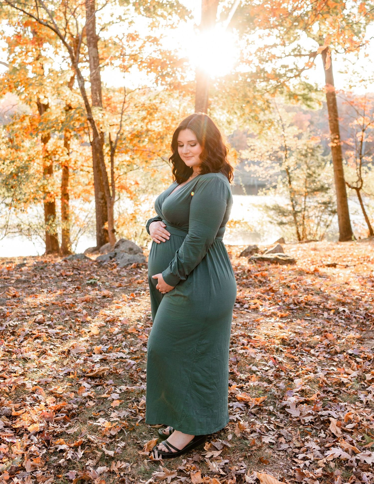 Golden sunset maternity photos with fallen leaves and lake backdrop.