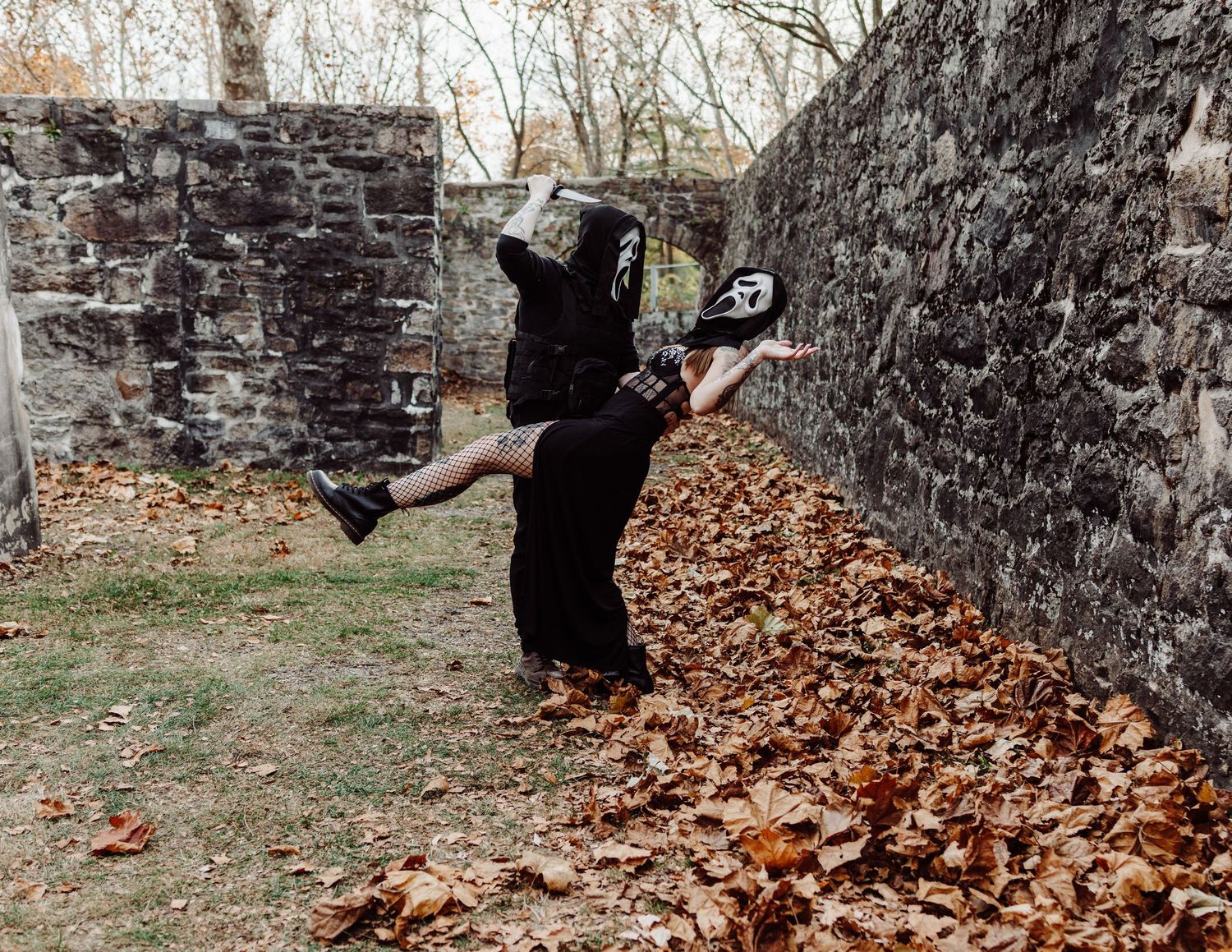Dark gothic photoshoot with fallen leaves and stone wall backdrop.