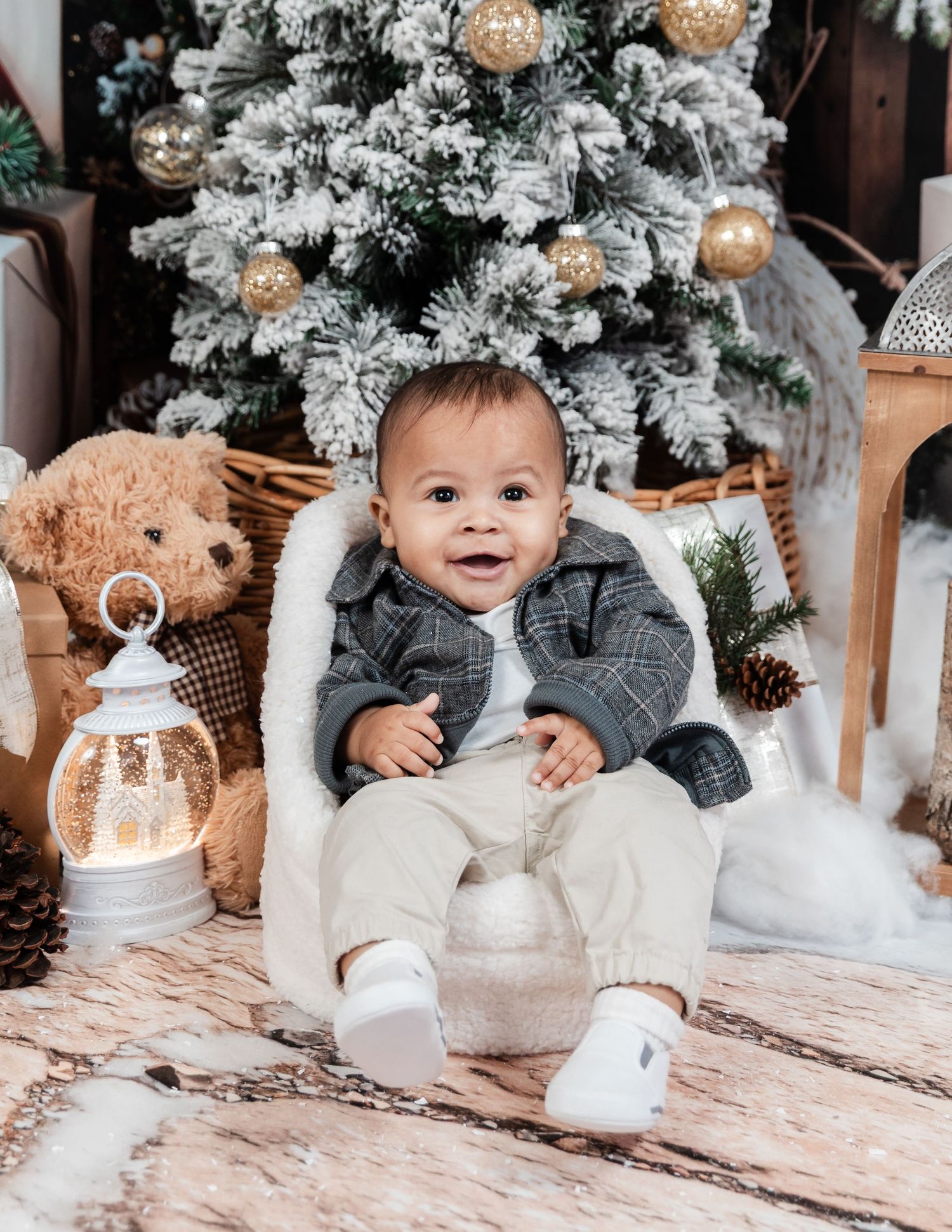 Baby sits on floor with milk bottles and teddy bear among snowy winter decorations.