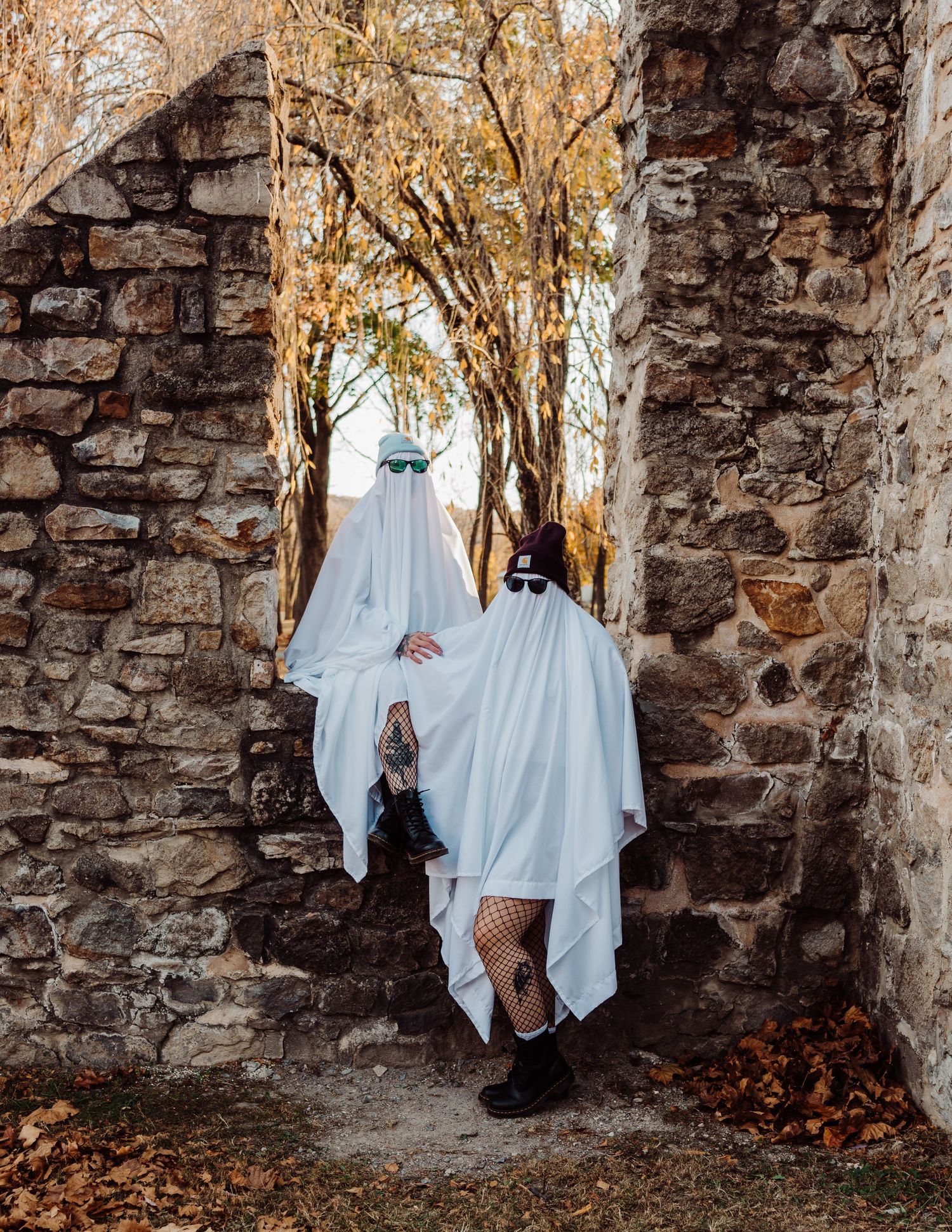 Ghost-costumed figures pose playfully against rustic stone walls in autumn setting.