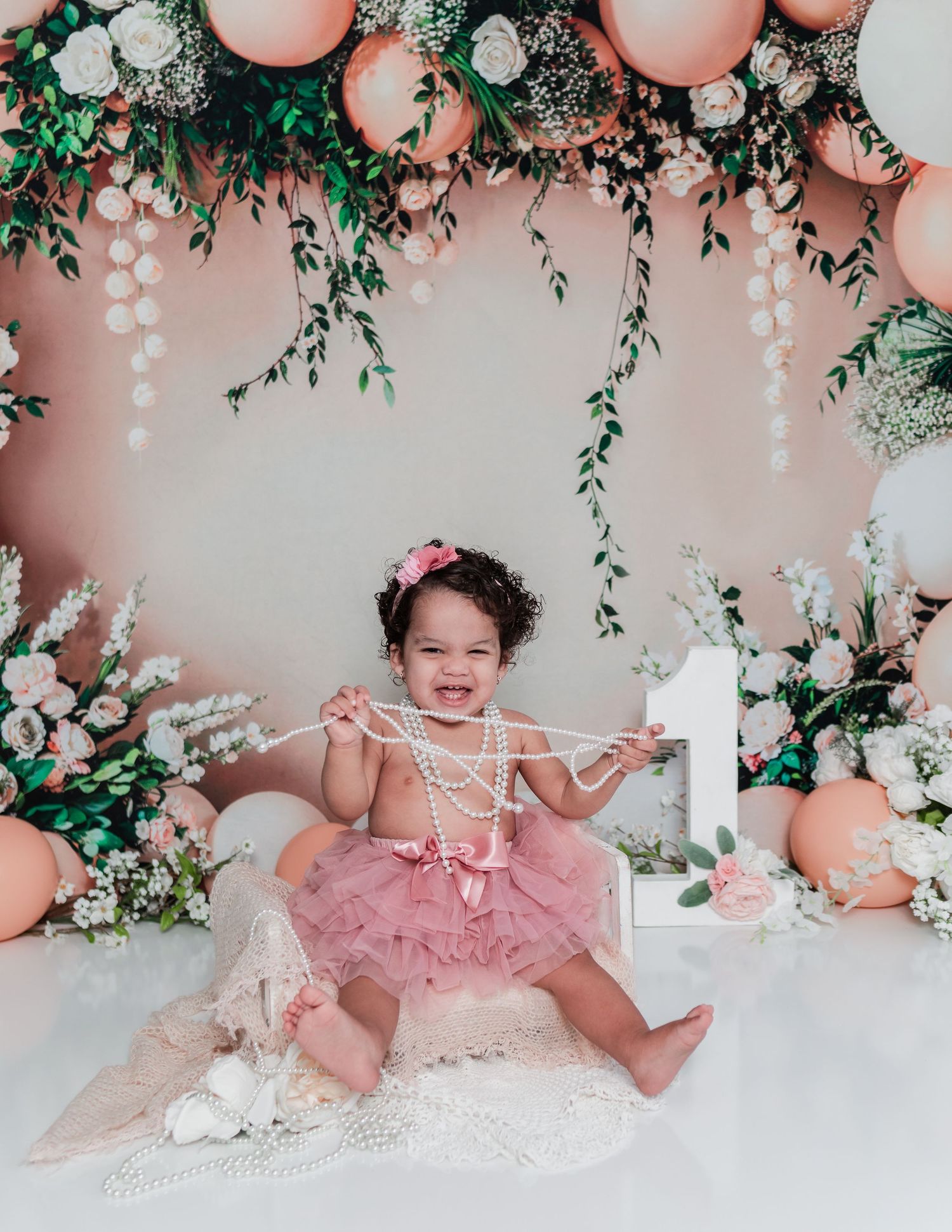 Baby girl in pink tulle dress at floral themed party with balloons and greenery decorations.