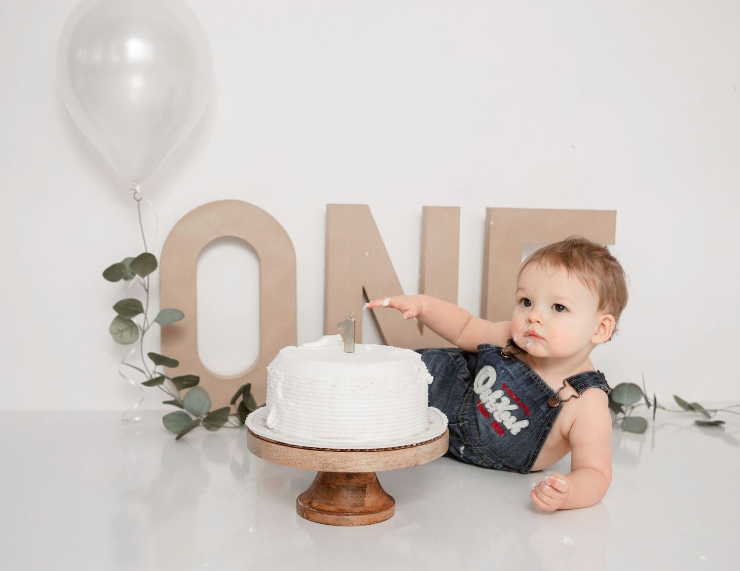 Minimalist cake smash setup with white balloon, eucalyptus, and wooden ONE letters.