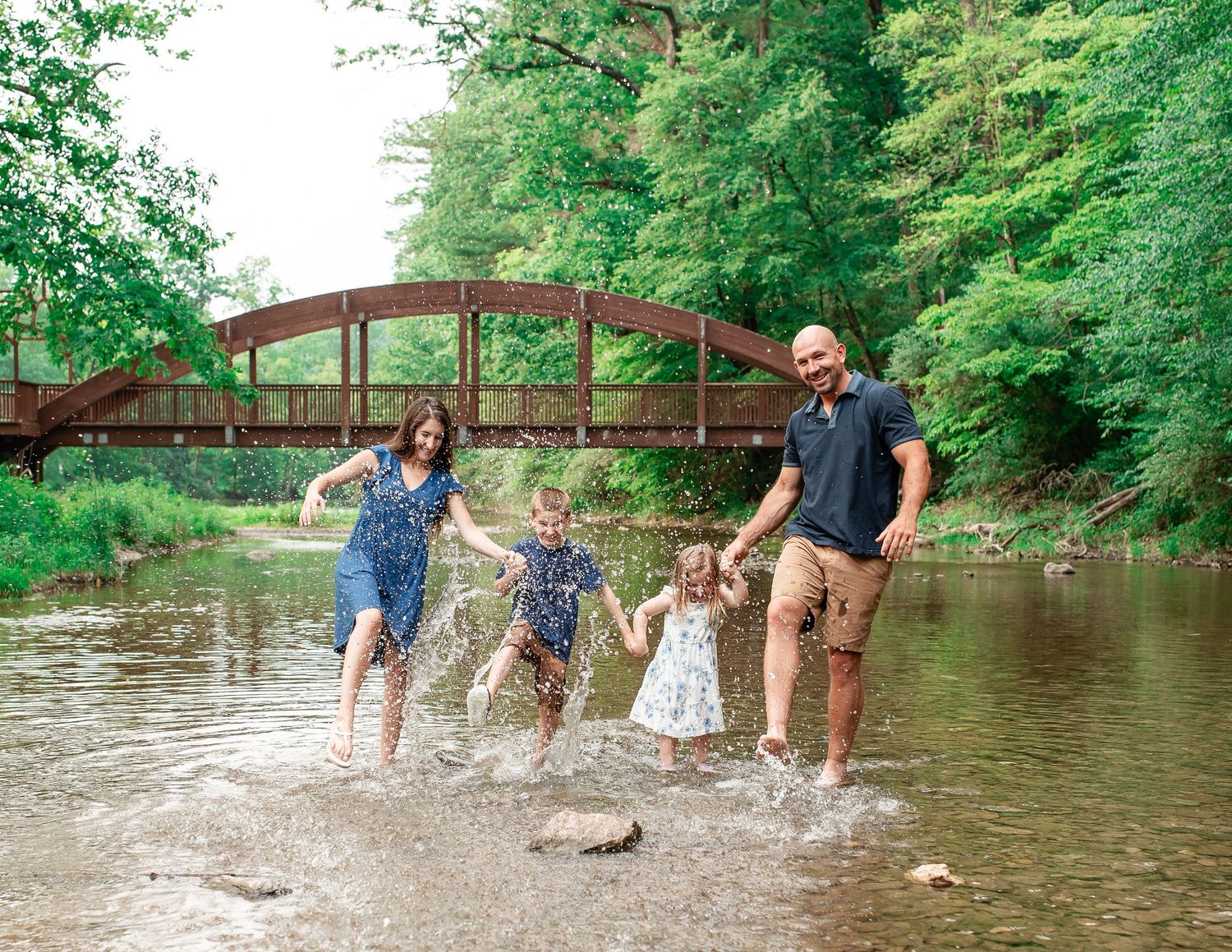 People splashing and playing in a shallow river near a rustic wooden bridge on a summer day.