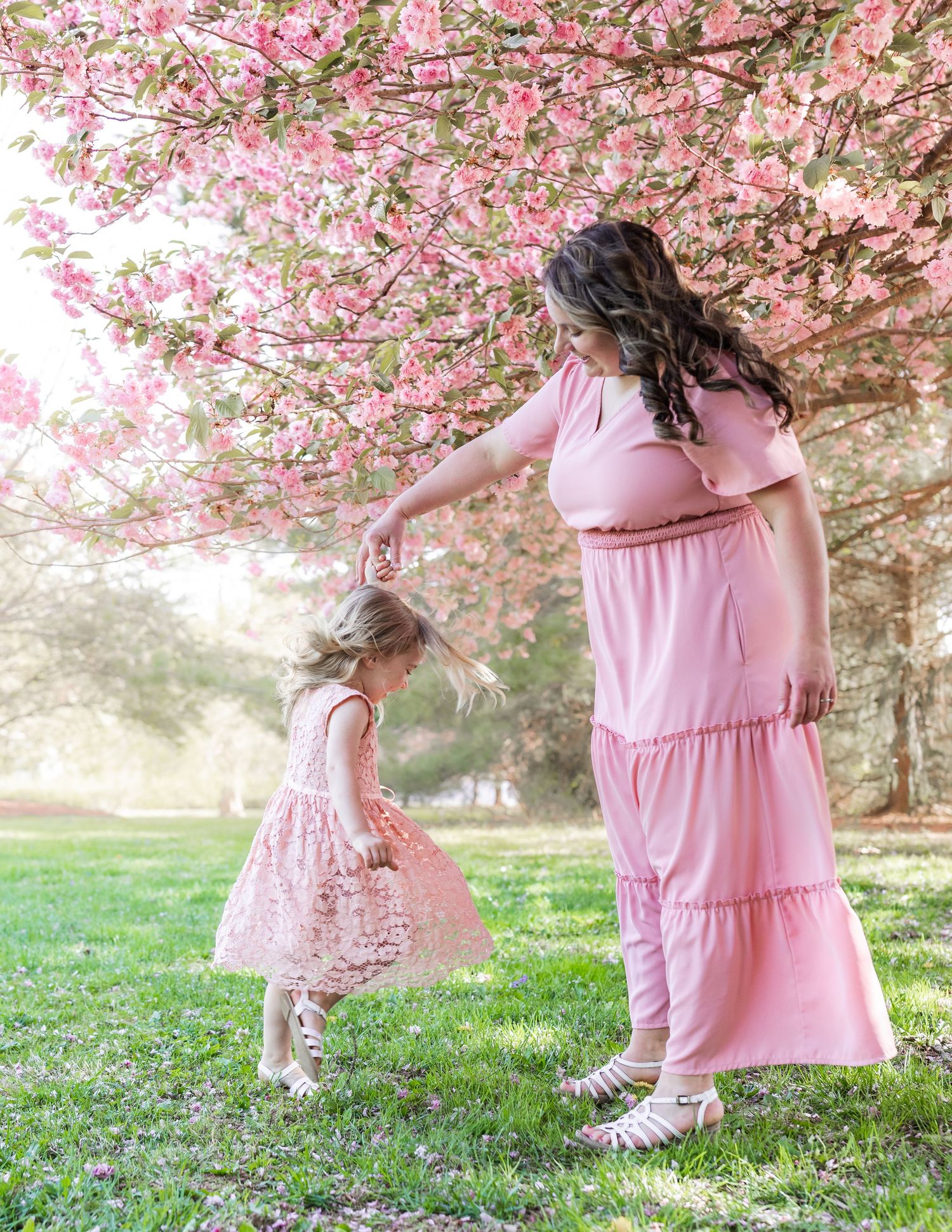 Pink-clad figures dance and twirl under a canopy of cherry blossoms in a dreamy spring setting.