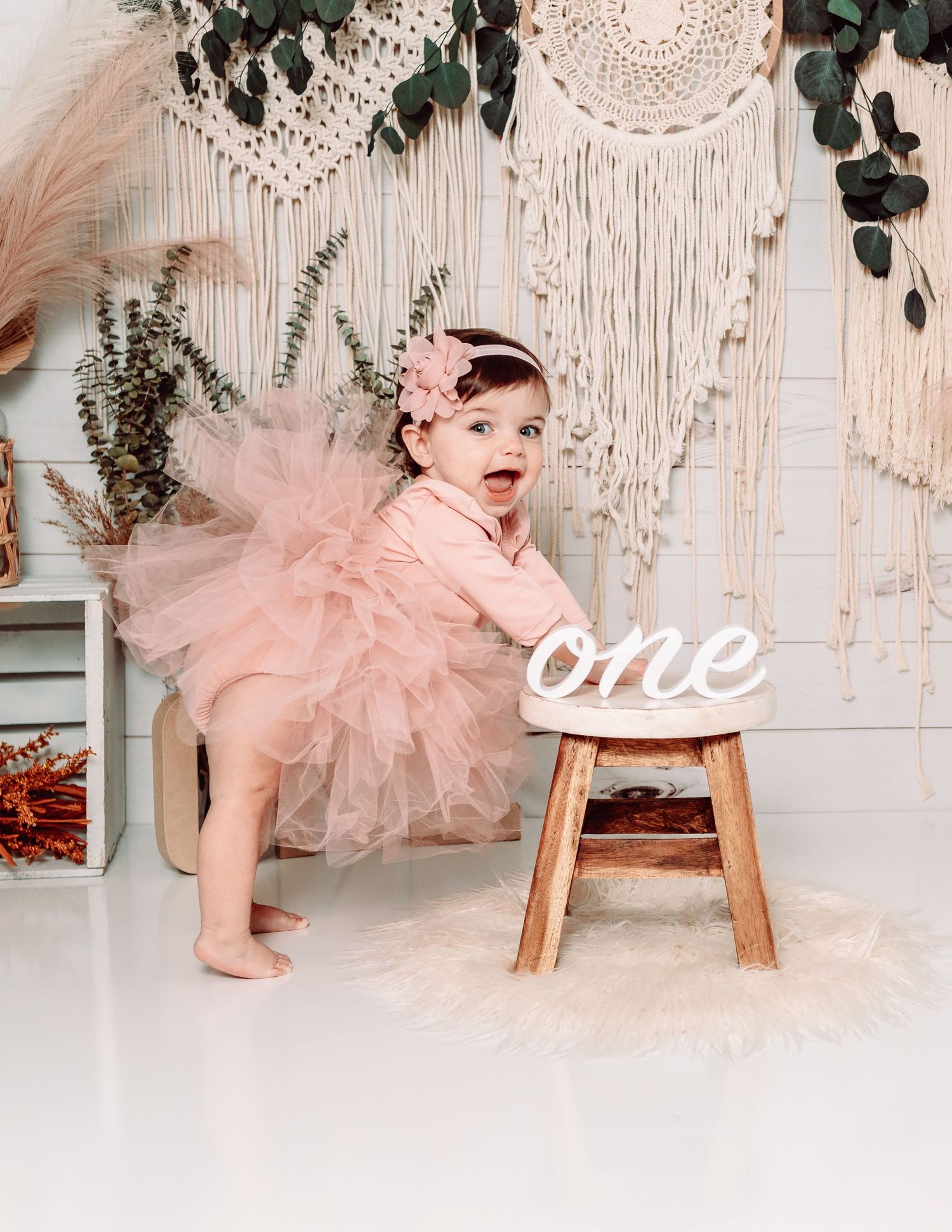 Baby in pink tulle dress celebrates first birthday with rustic boho decorations and macrame backdrop.