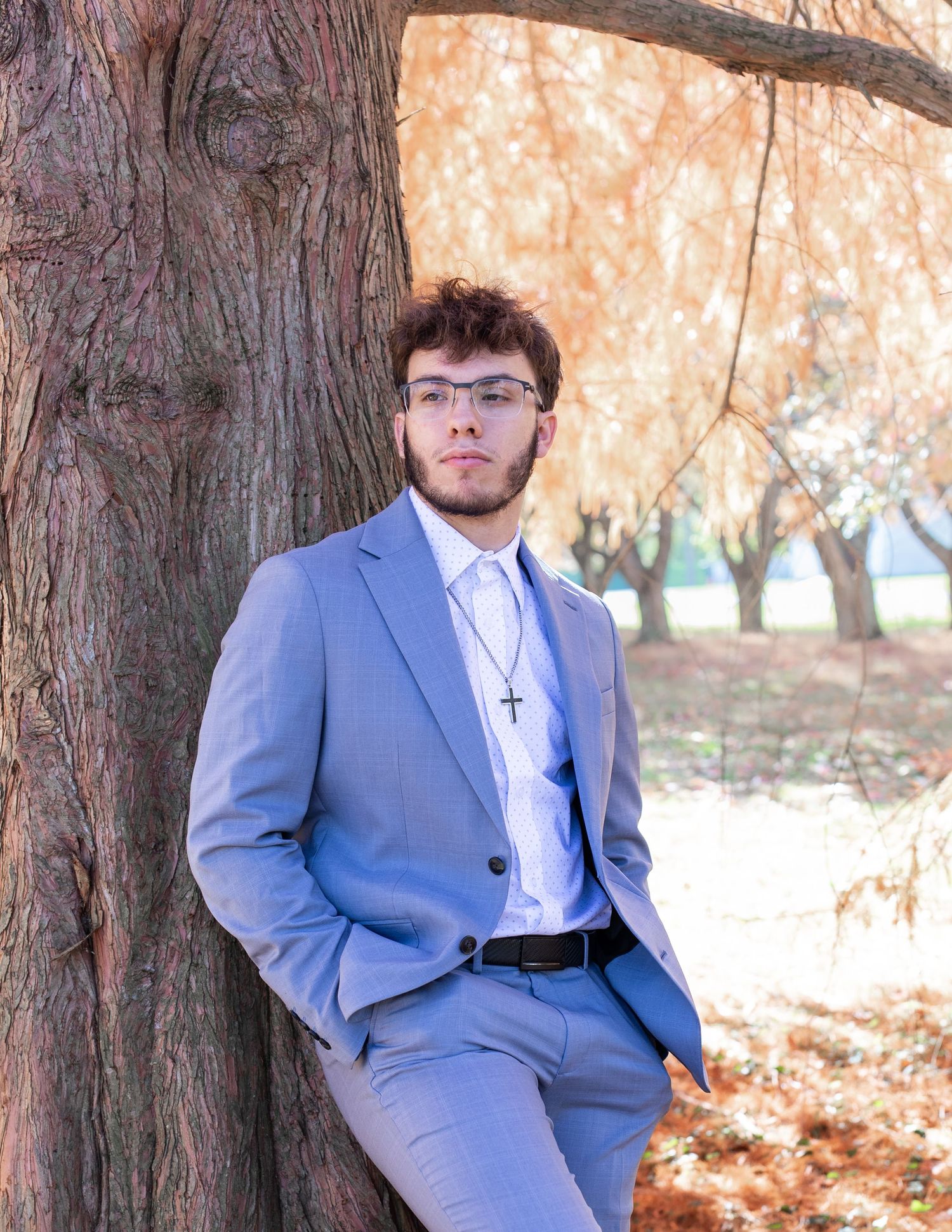 Person in blue suit leans against tree trunk with autumn leaves in background.