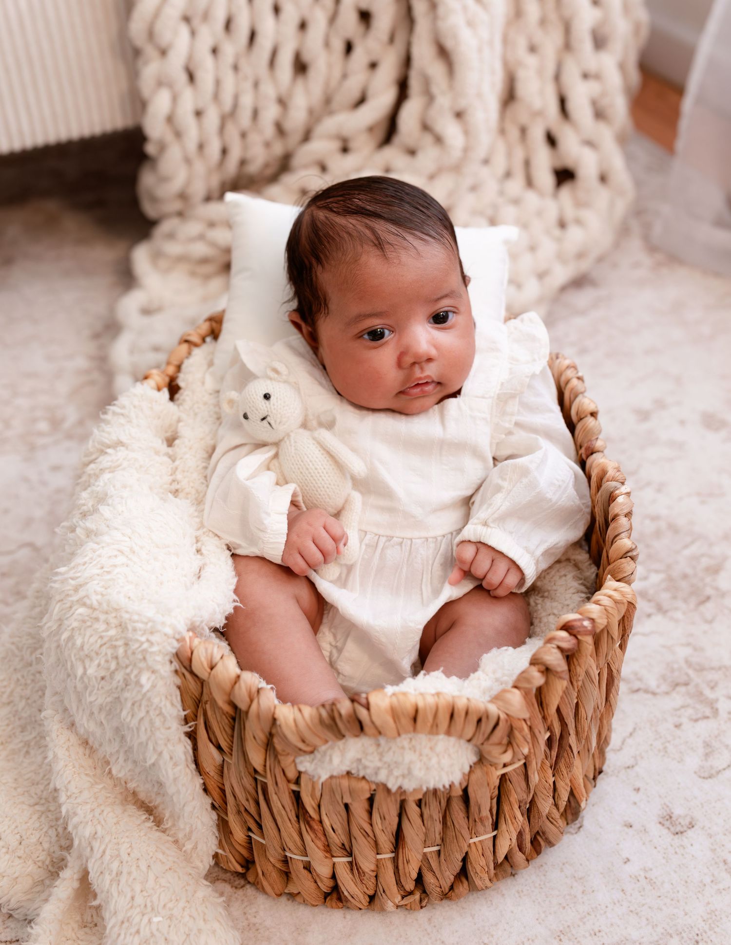 Small baby wearing white outfit sits in woven basket lined with soft sheepskin against chunky knit blanket backdrop.