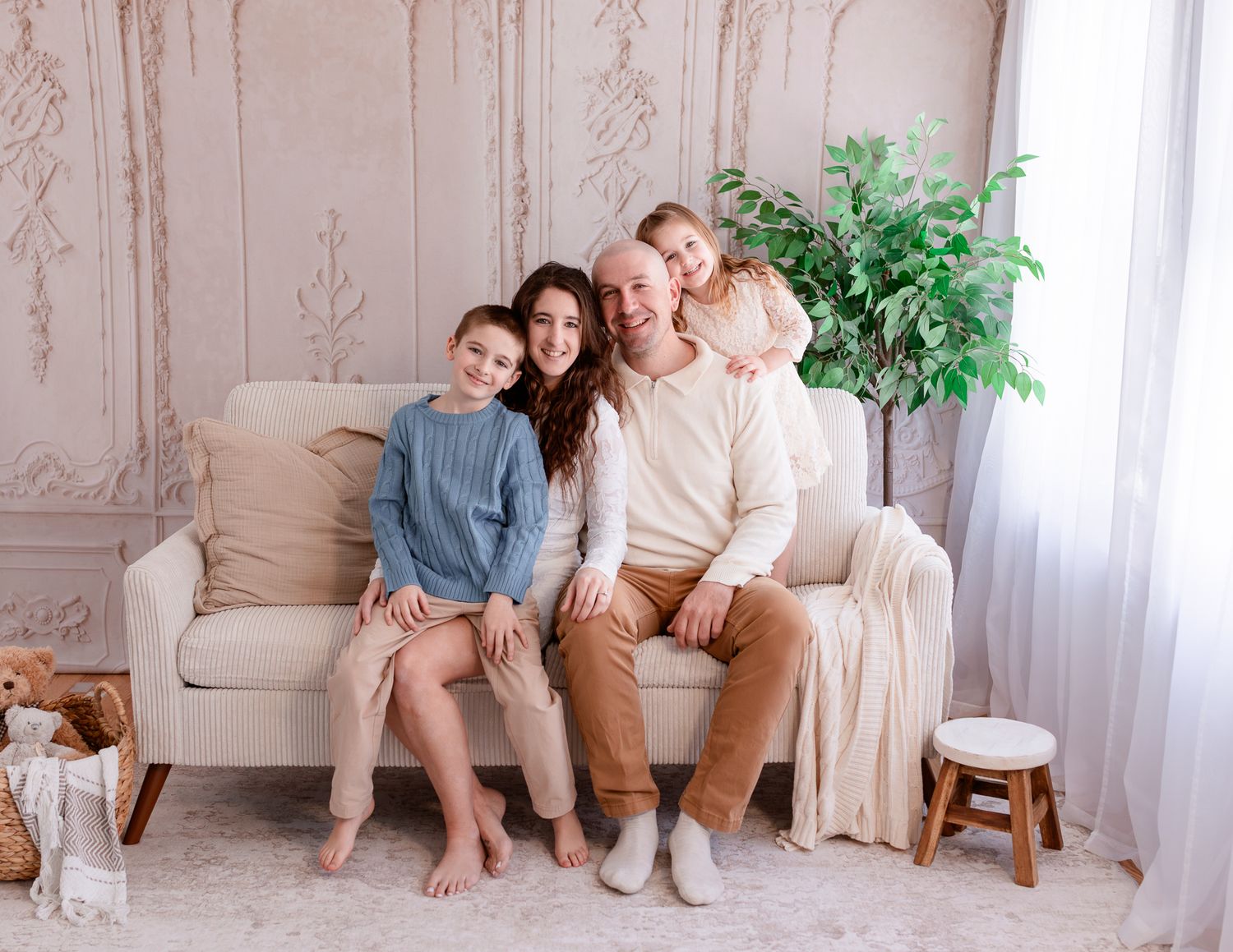 A family sits together on a beige couch in a bright room with decorative wall panels and a potted plant.
