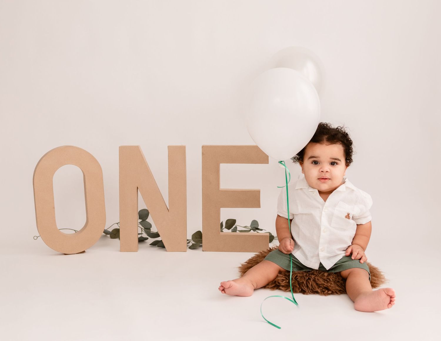 A baby in a white onesie sits on a white backdrop with tan colored wooden letters displayed behind them.