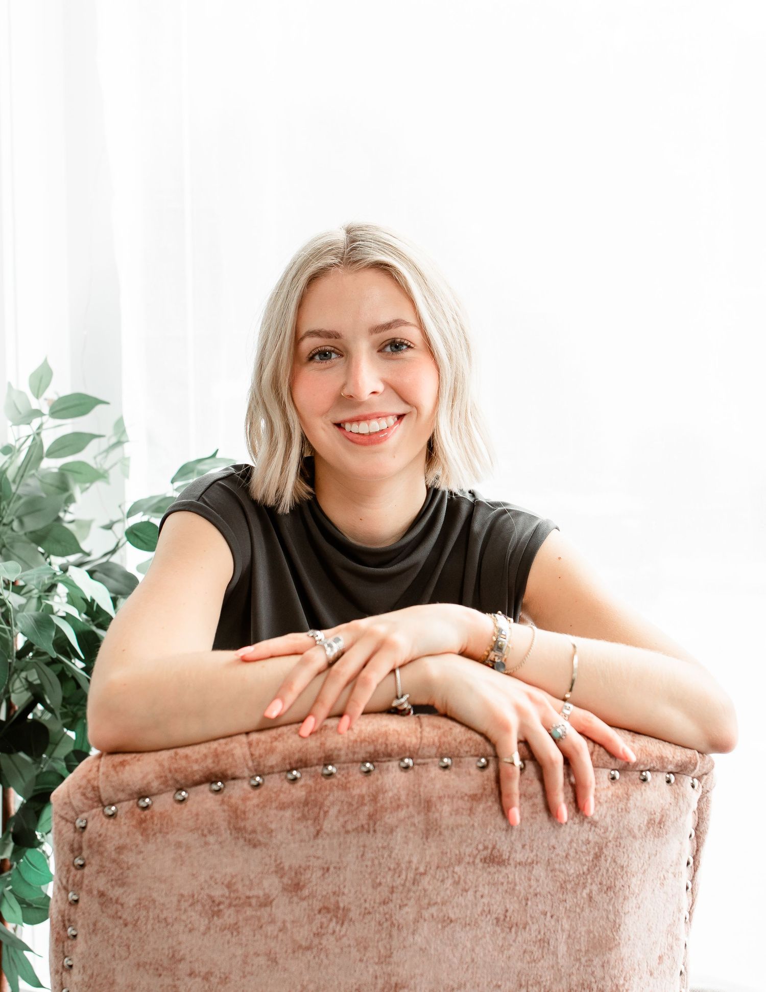 Woman in black top leans on velvet chair with plant in bright setting.